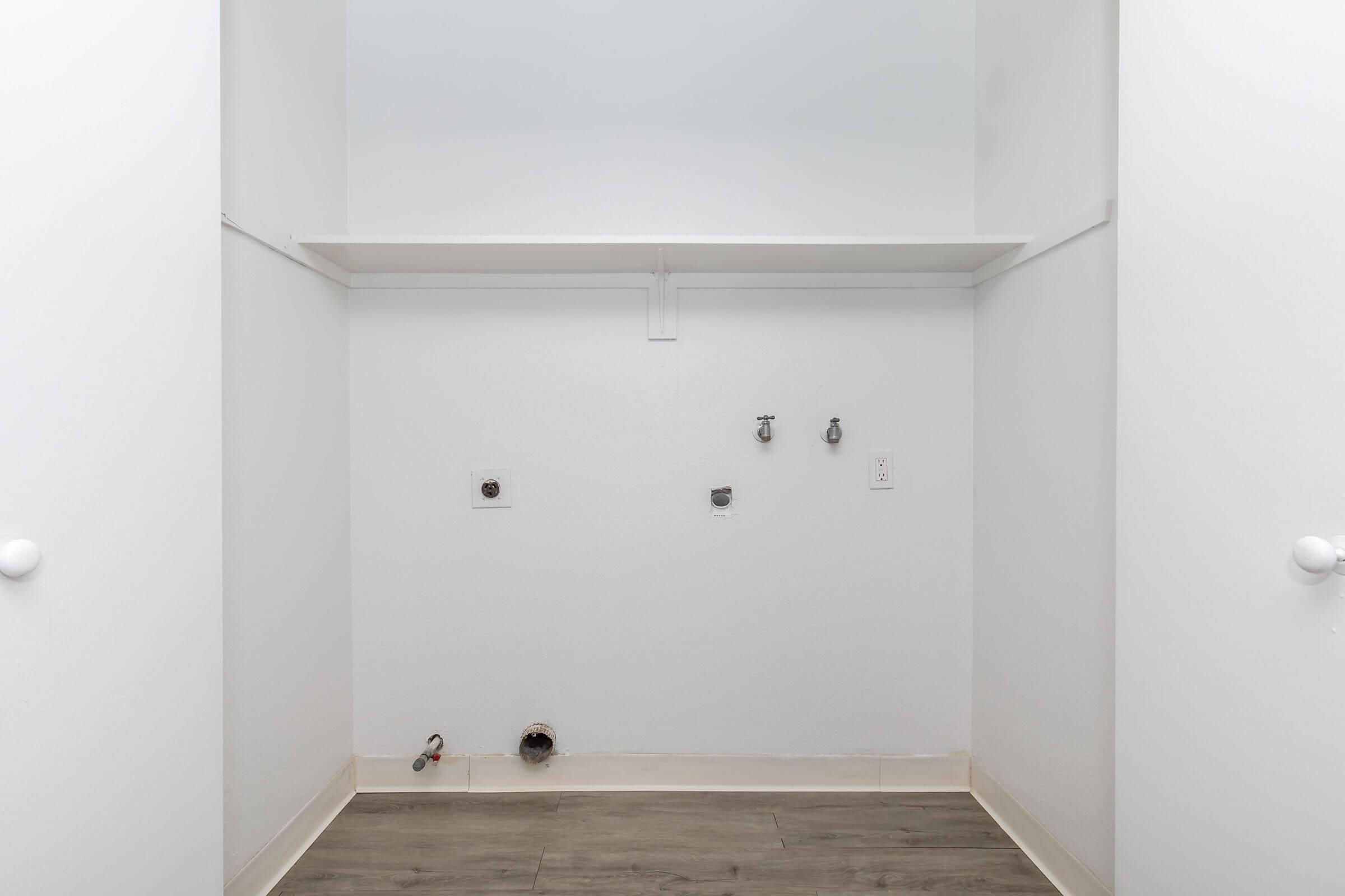 Empty laundry room with a clean, white interior. A shelf is mounted against the wall, and there are two water connectors and an electrical outlet visible. The floor is a light-colored laminate, and the overall space is bright and minimalist.