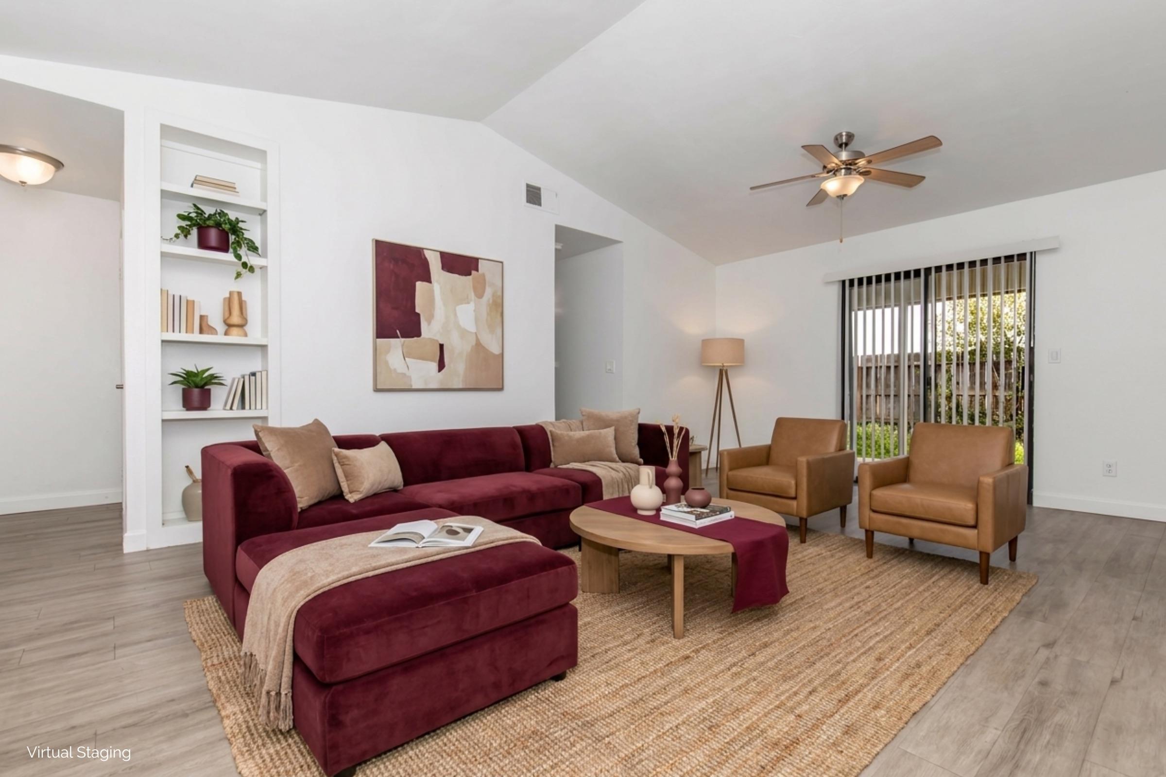 A modern living room featuring a burgundy sectional sofa with accent pillows, two brown armchairs, a round coffee table, and a decorative area rug. The walls are white with a neutral artwork piece. There are shelves displaying plants and books, and a ceiling fan above. Natural light filters in through sliding glass doors.