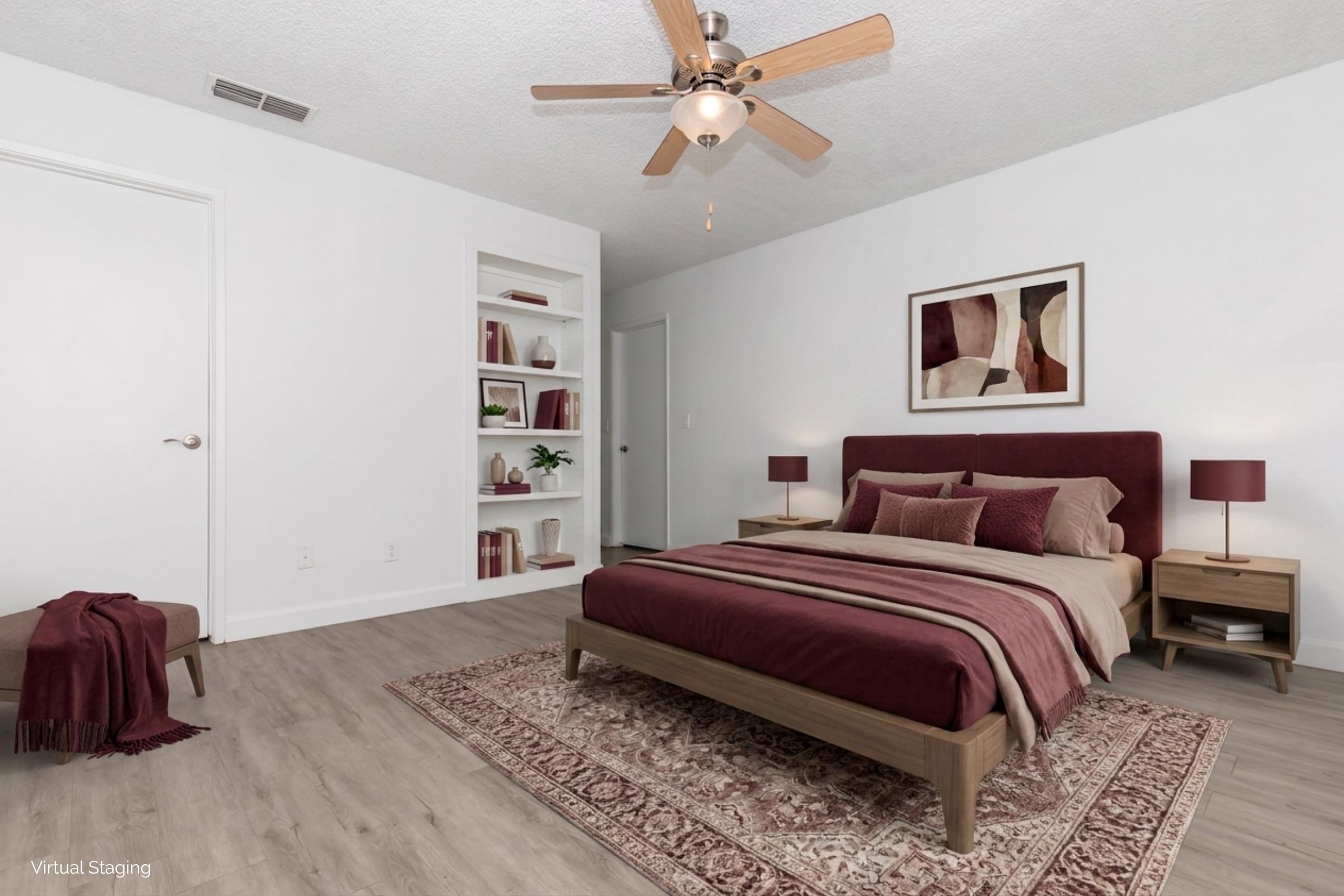 A modern bedroom featuring a queen-sized bed with burgundy bedding, matching nightstands, and lamps. A wall-mounted artwork adds color, while a small bookshelf showcases decorative items. The room has light wood flooring and a warm atmosphere, enhanced by a ceiling fan.