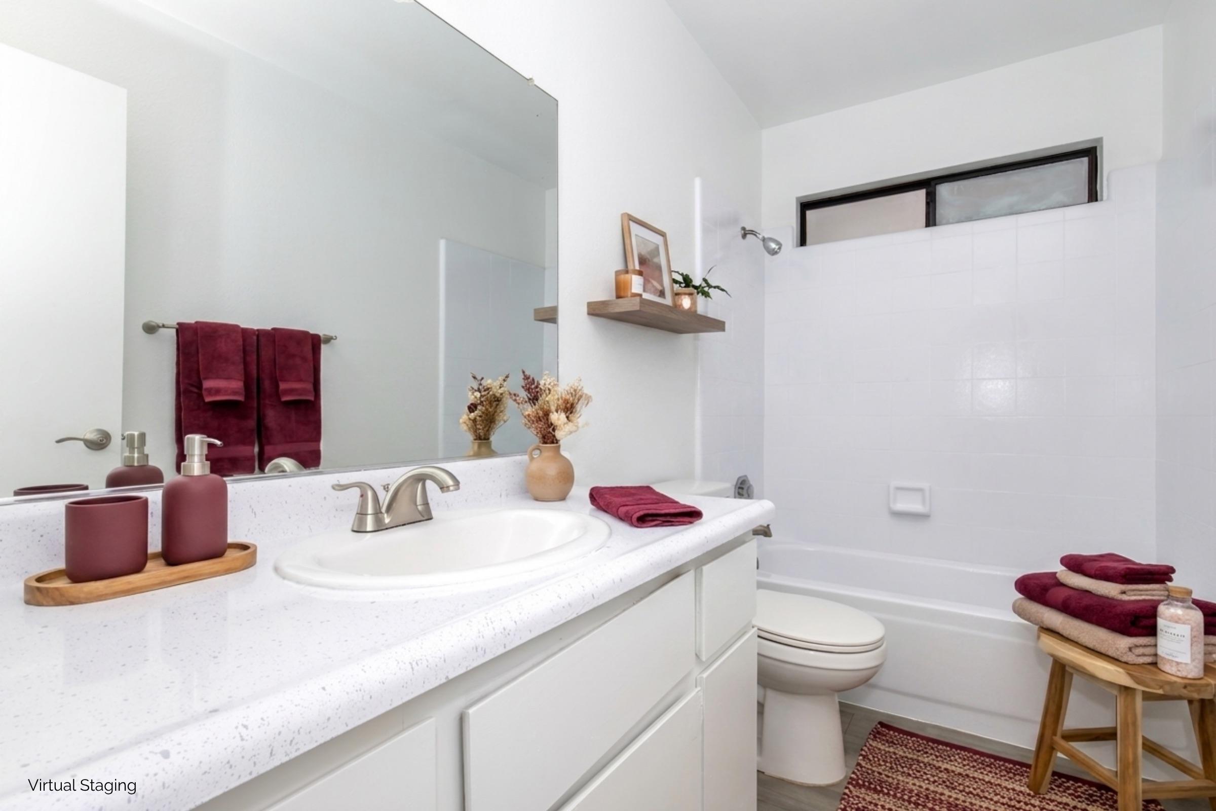 A clean, modern bathroom featuring a white countertop with decorative items, a large mirror, and a minimalist design. The shower area is enclosed with white tiles. Towels in deep burgundy hang on the wall, complemented by a wooden shelf with small plants and accessories, creating a warm and inviting atmosphere.
