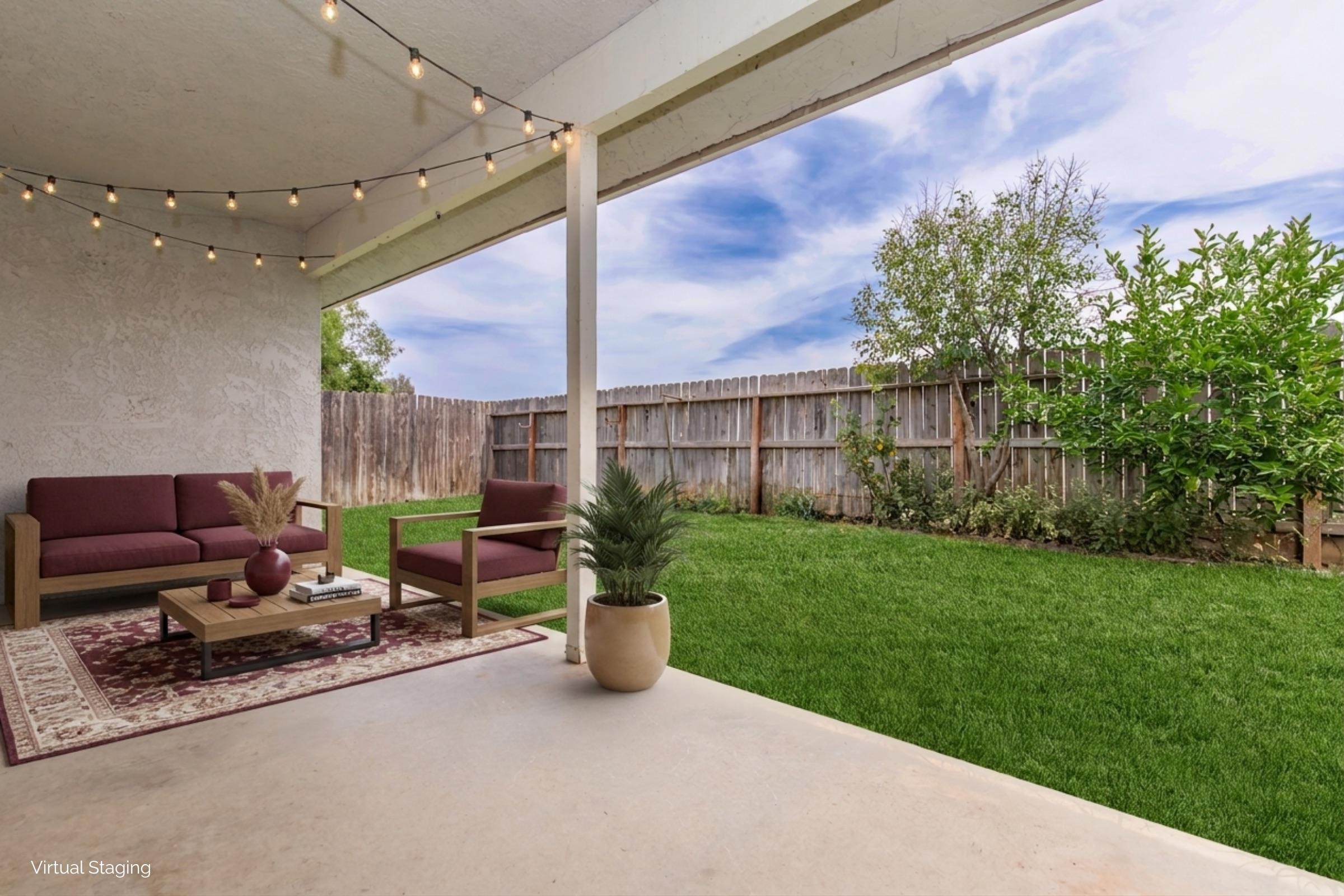A cozy outdoor patio area featuring a small seating arrangement with two chairs and a coffee table. The space is adorned with string lights overhead. In the background, there's a well-maintained grassy yard bordered by a wooden fence and lush shrubs. The atmosphere is inviting and tranquil.