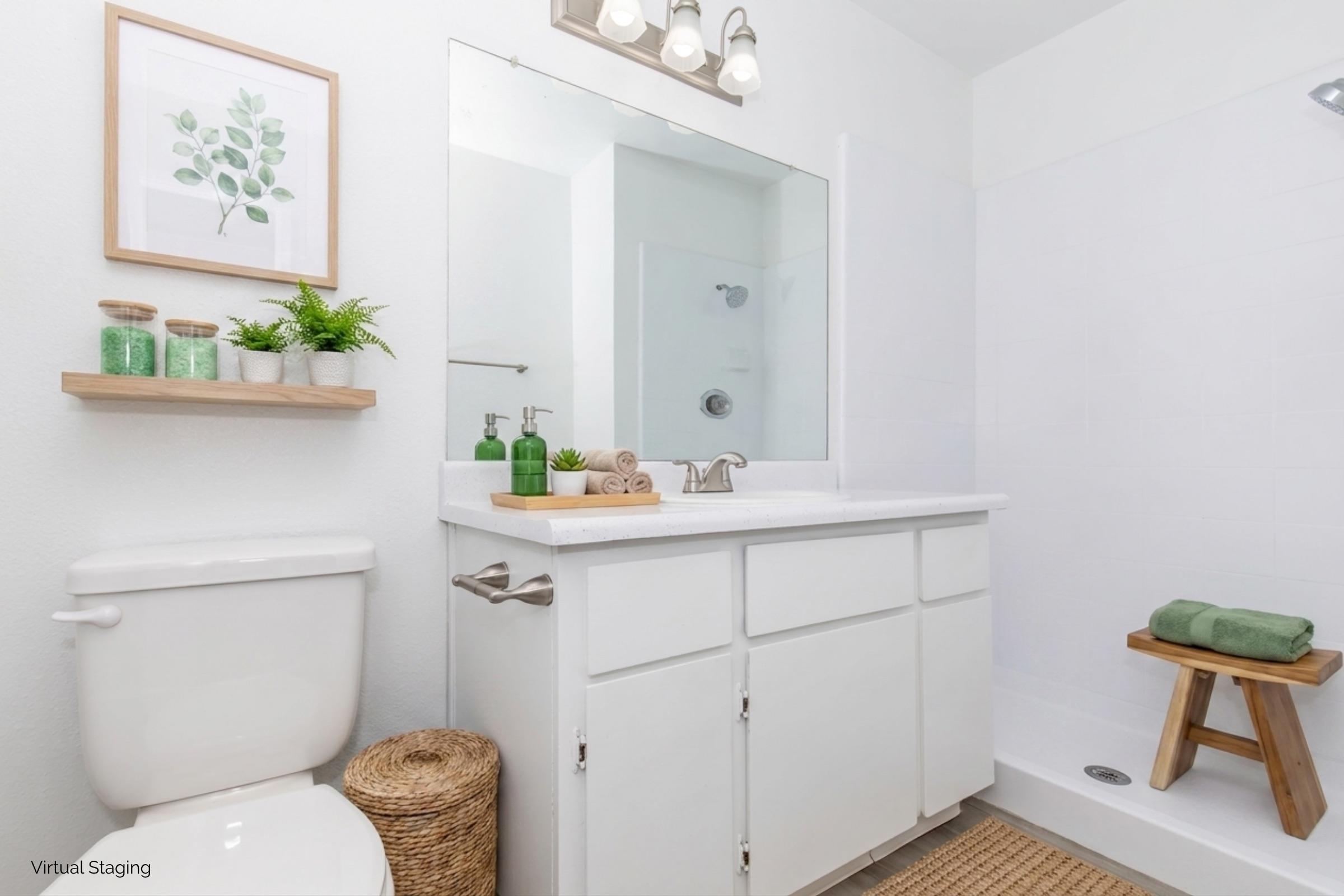 A modern bathroom featuring a white vanity with a large mirror, a toilet, and a shower. Shelves above the sink hold decorative plants and green toiletries. A wooden stool and towels are neatly arranged, while a woven rug adds warmth to the space. Soft, neutral colors create a calming atmosphere.