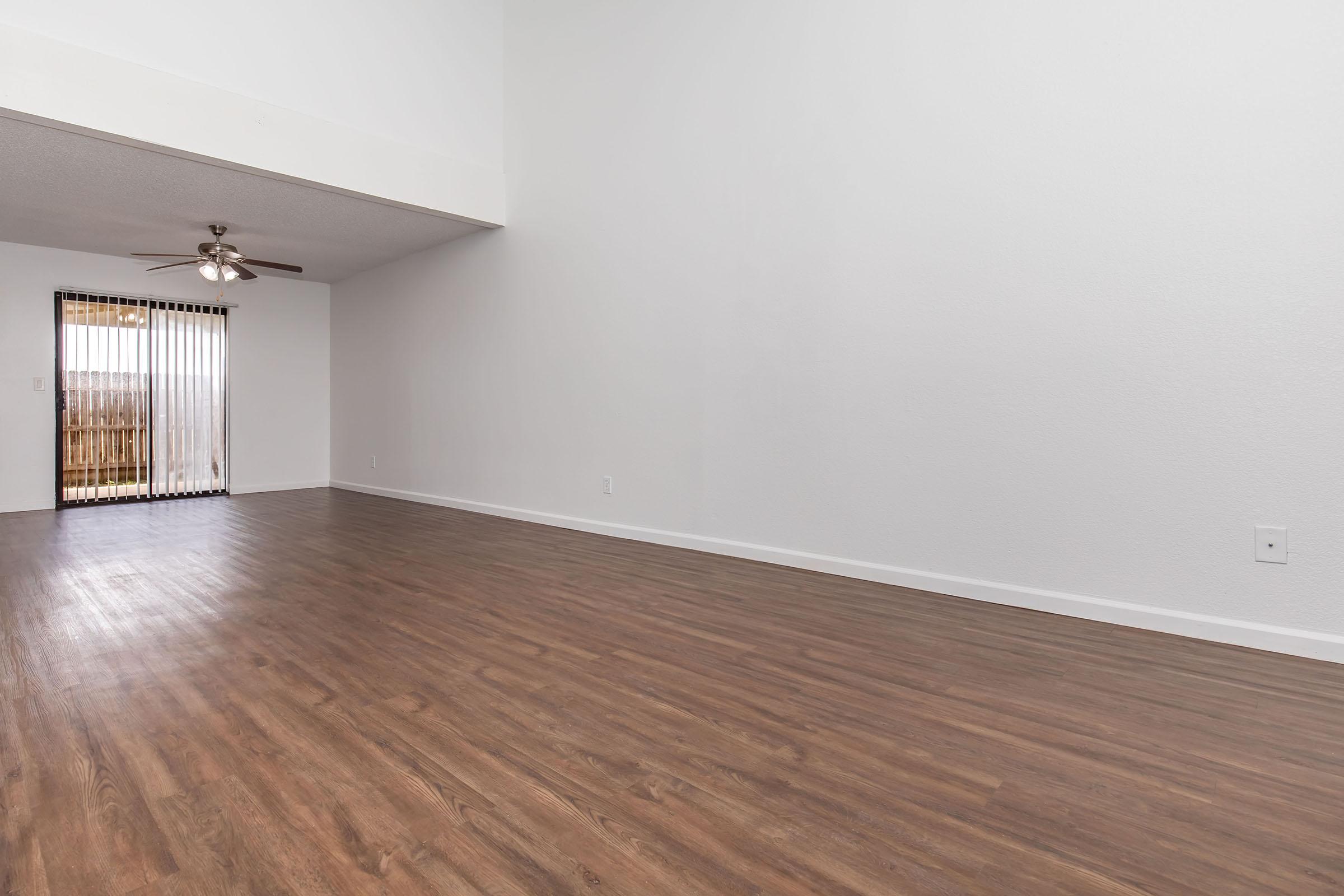 A spacious, empty living room with wooden flooring, a ceiling fan, and white walls. Large windows with vertical blinds allow natural light to enter, and a sliding glass door leads to an outdoor area. The high ceiling creates an open and airy atmosphere.