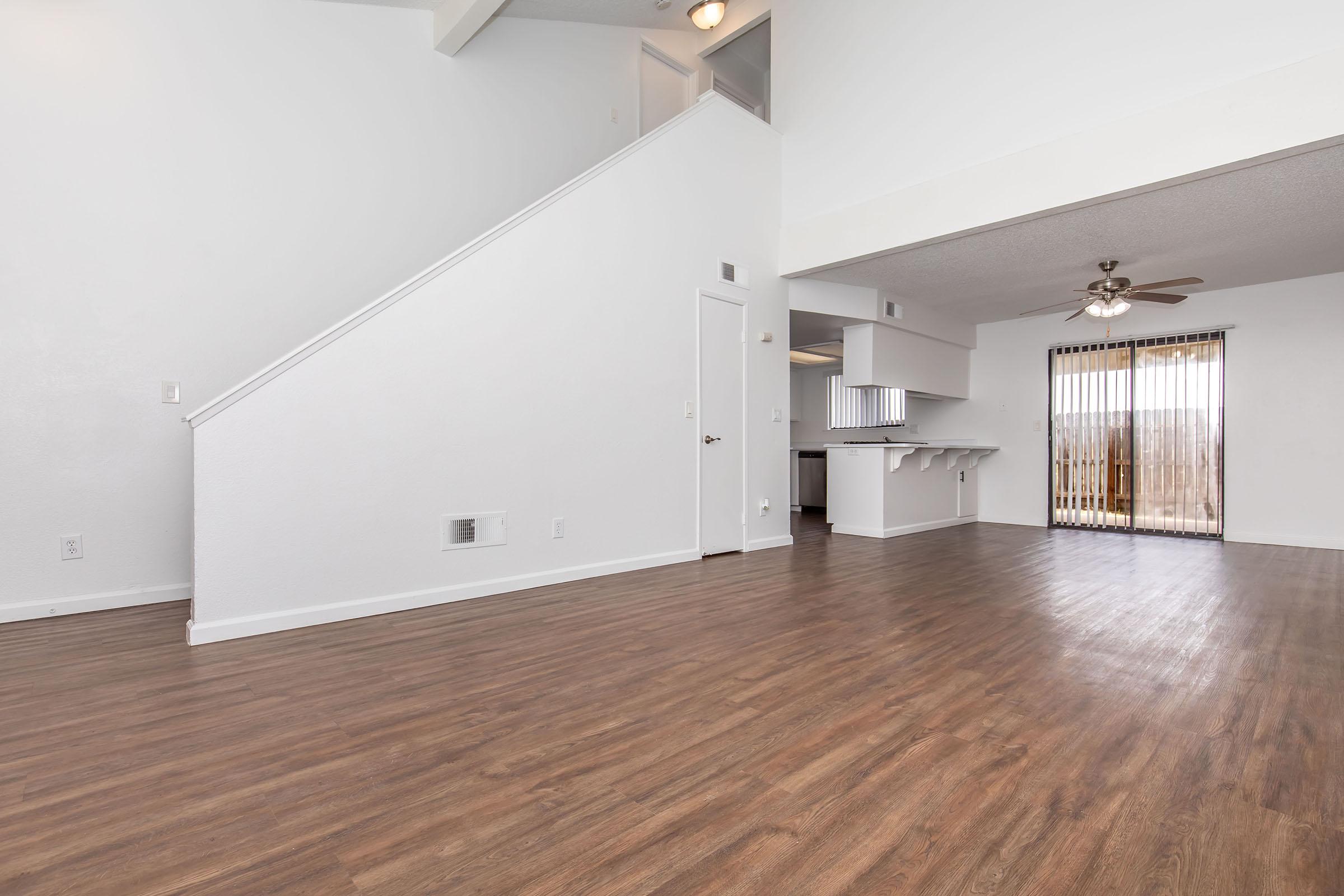 Spacious living area with hardwood floors, white walls, and a high ceiling. Features an open layout with a staircase leading to an upper level. Includes sliding glass doors that open to a balcony or patio area, providing natural light. Kitchen area visible in the background.