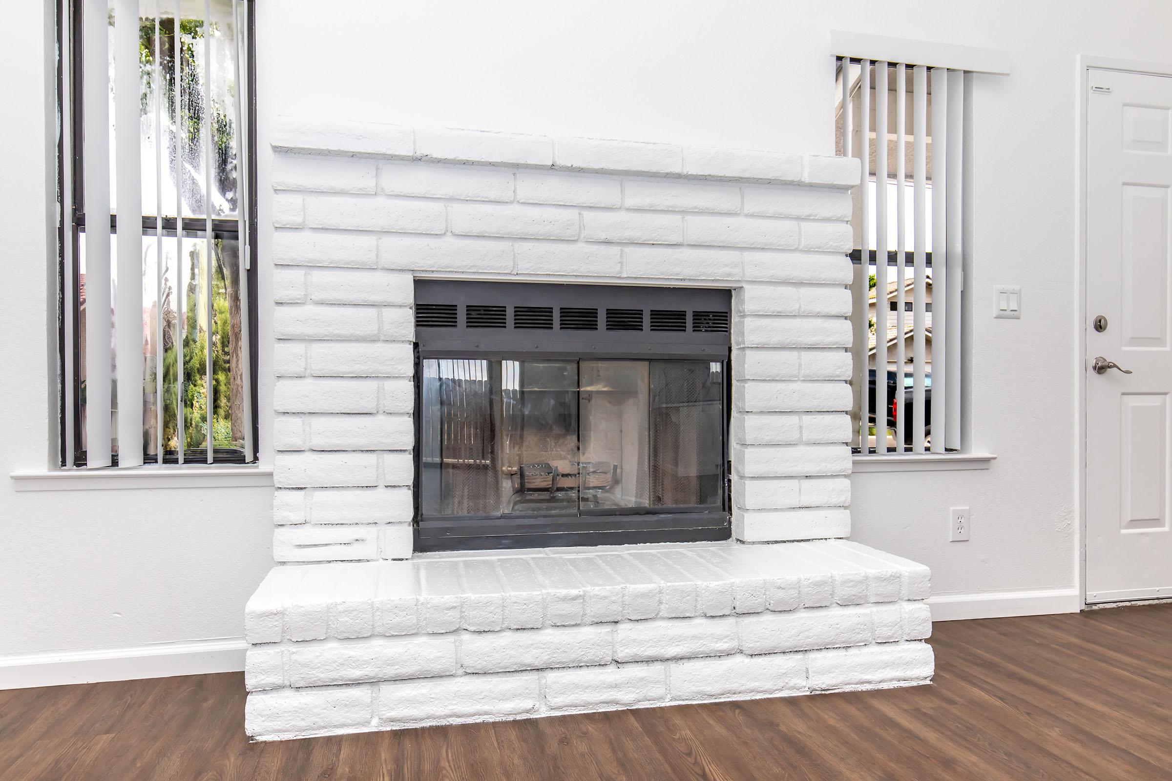 A white brick fireplace with a glass front is the focal point of a modern living room. The wall surrounding the fireplace is painted white, and there are vertical blinds on two windows, allowing natural light to enter. The floor features dark wood laminate, enhancing the contemporary aesthetic.