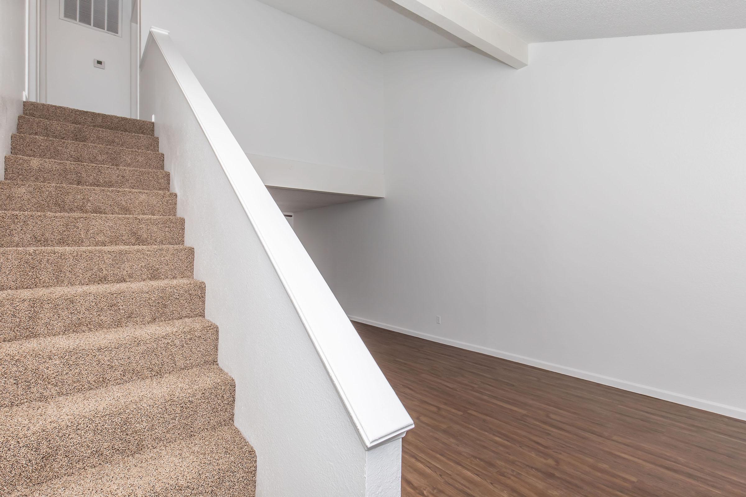 Interior of a modern home showcasing a staircase with beige carpet leading to a landing. The walls are painted white, and the floor features light brown laminate. The space is bright and minimalist, with a door visible at the top of the stairs and an open area on the lower level.