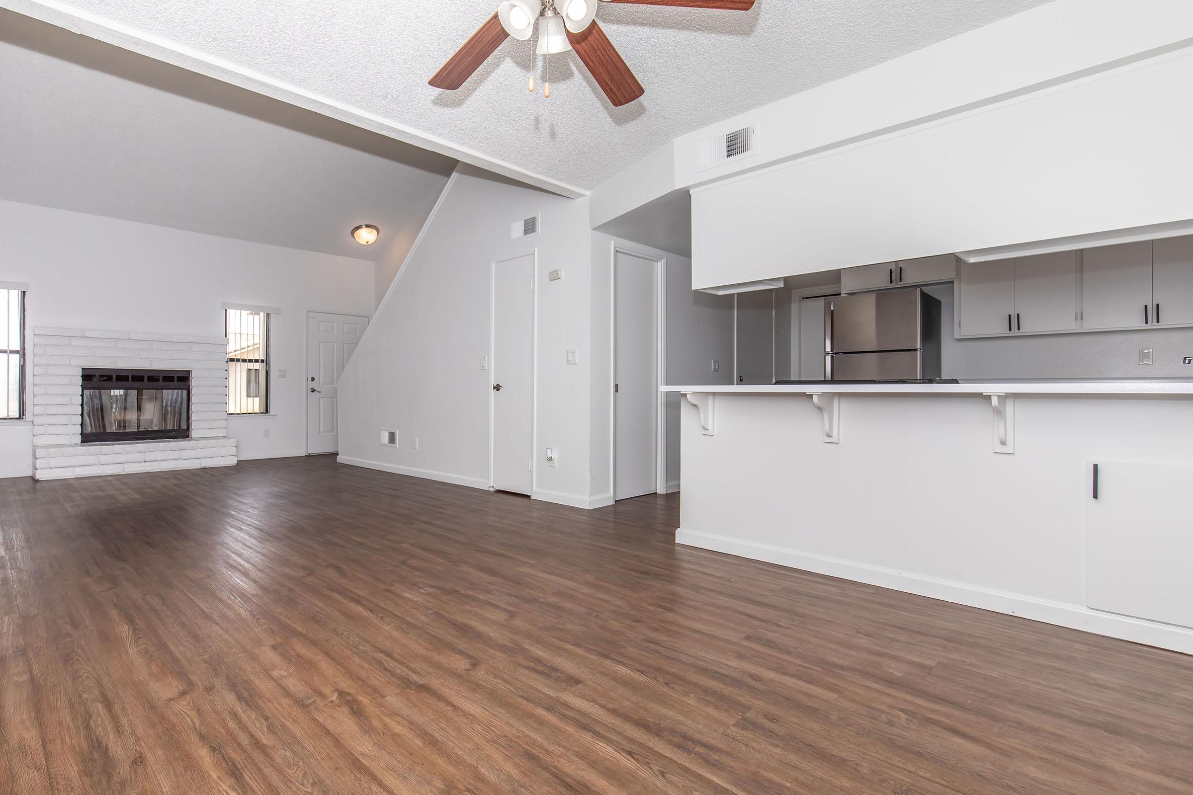 A spacious living room with hardwood-style flooring, featuring a ceiling fan and a fireplace. The room has three large windows allowing natural light, with a doorway leading to another area of the house. The walls are painted white, and the kitchen area is partially visible in the background with modern cabinetry.