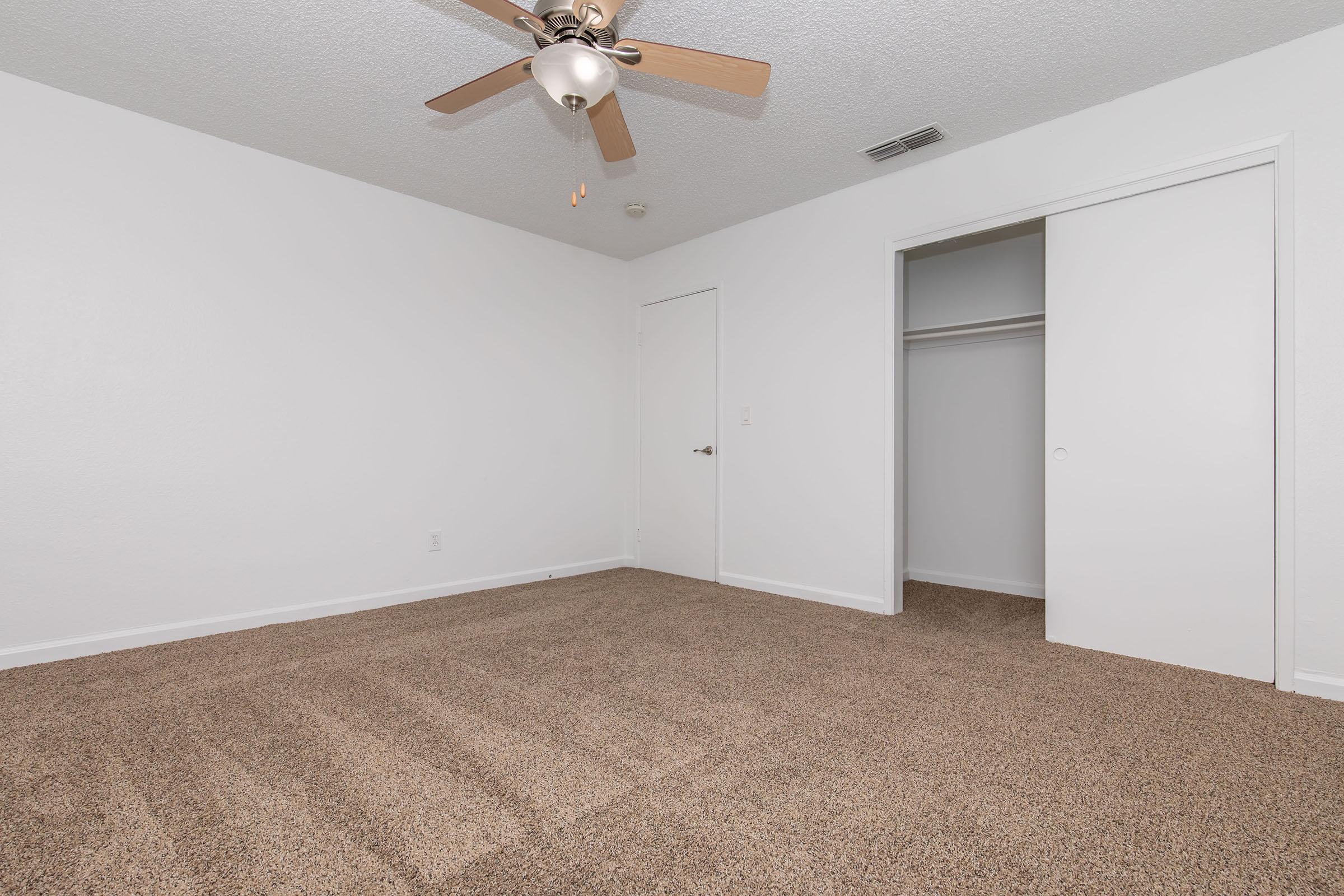 A spacious room with light-colored walls and a ceiling fan. The floor is covered in brown carpet. There is a closet with sliding doors on one side, and a plain white door leading to another area. Natural light may illuminate the room, creating a bright atmosphere.