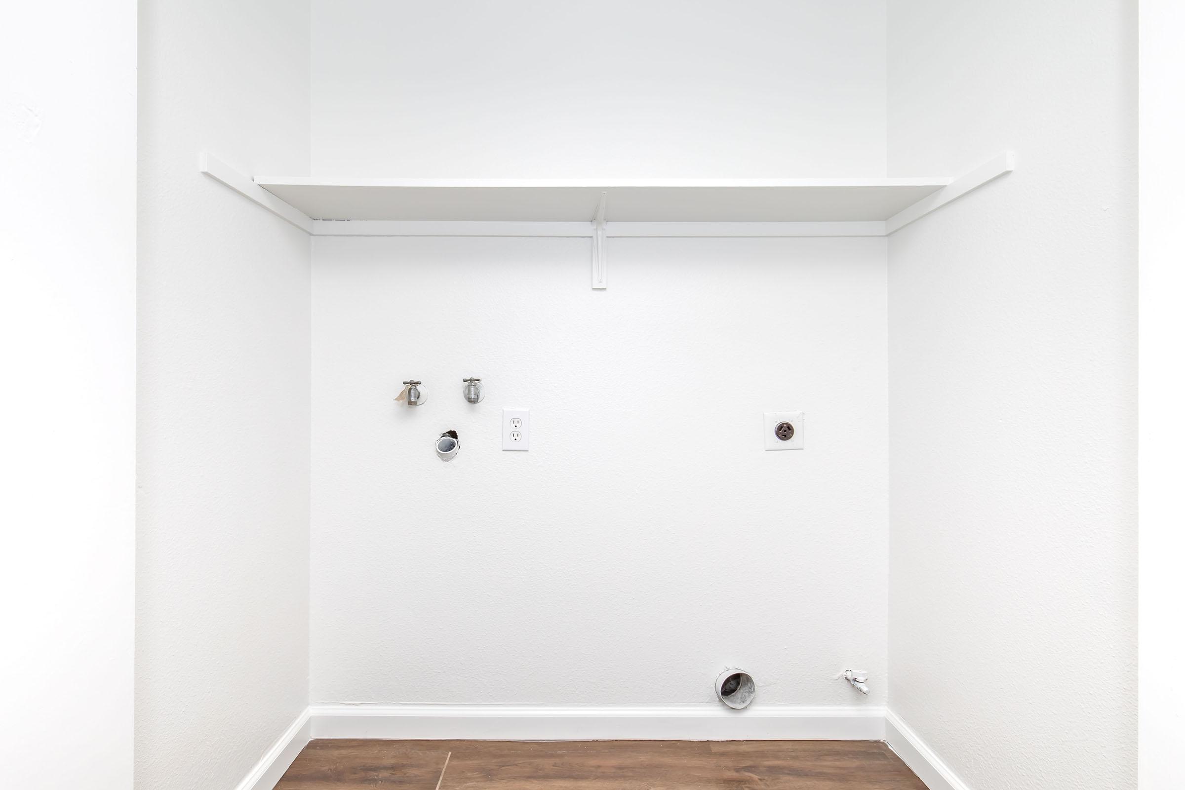 Empty laundry room with a white wall. Features include a shelf above, water connections on the left, a power outlet, and a dryer vent on the right. The floor is made of brown wood-like material, creating a clean and minimalistic appearance.