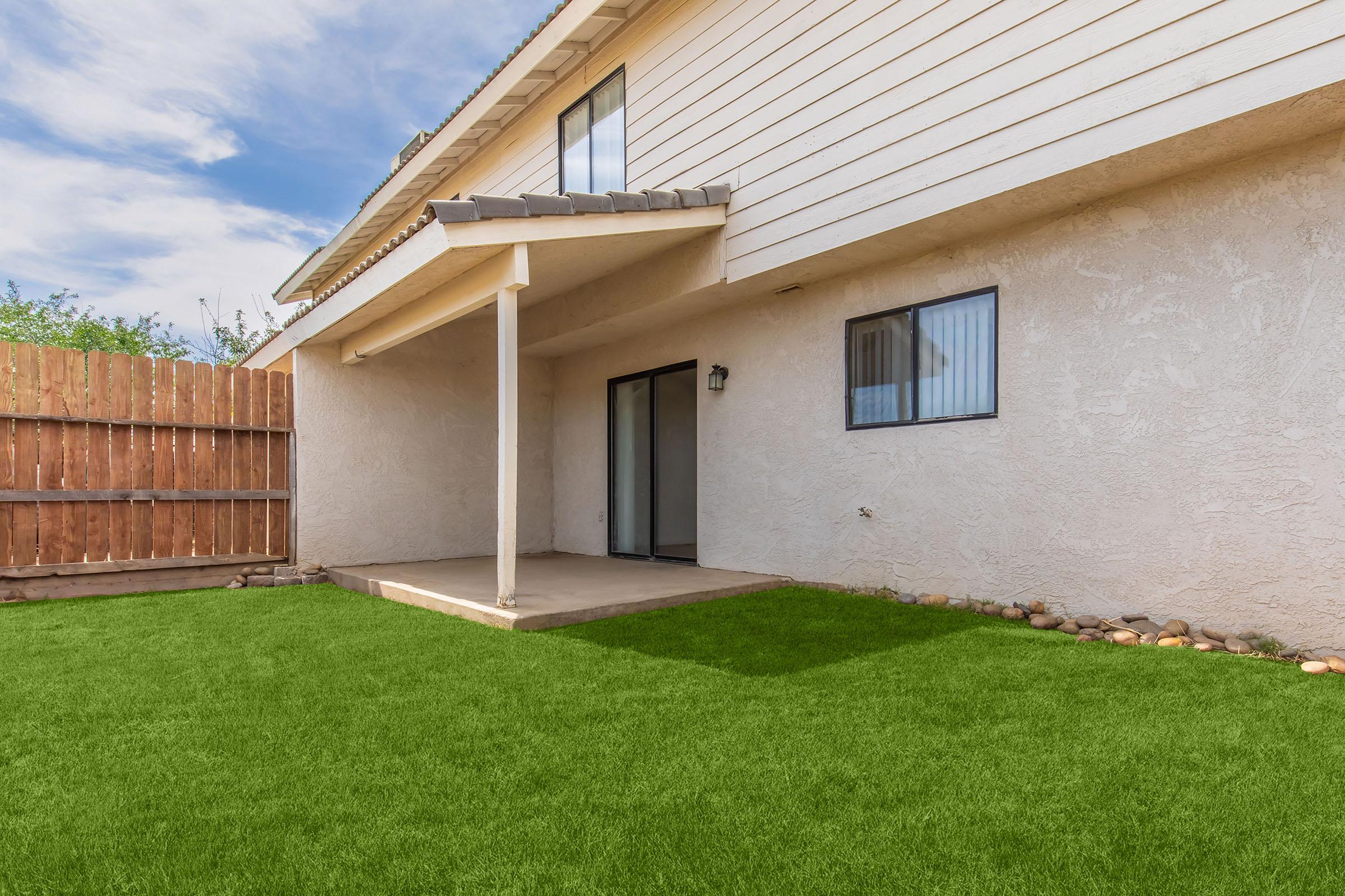 A view of the back patio of a house featuring a well-maintained lawn with artificial grass. The patio, covered by a roof, has sliding glass doors and a single exterior light fixture. A wooden fence borders the yard, and the sky is partly cloudy, creating a bright outdoor atmosphere.