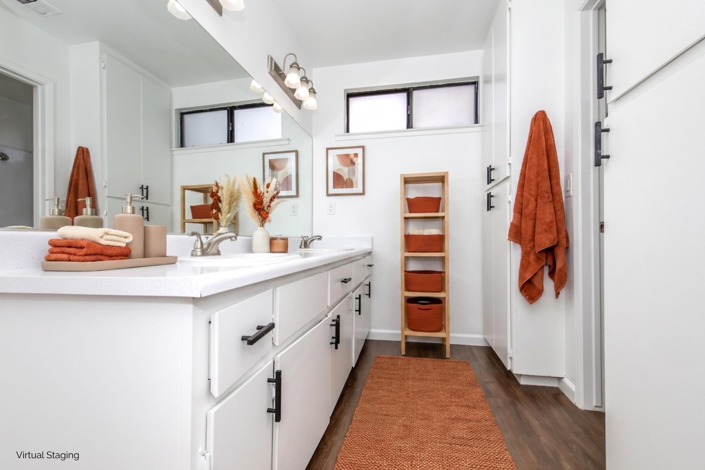 A modern bathroom featuring a double sink vanity with white countertops, framed mirror, and wood accents. Orange towels hang on a rack, and a decorative arrangement with orange hues complements the decor. A light natural wood floor adds warmth, while a fabric runner leads to a small storage unit nearby.