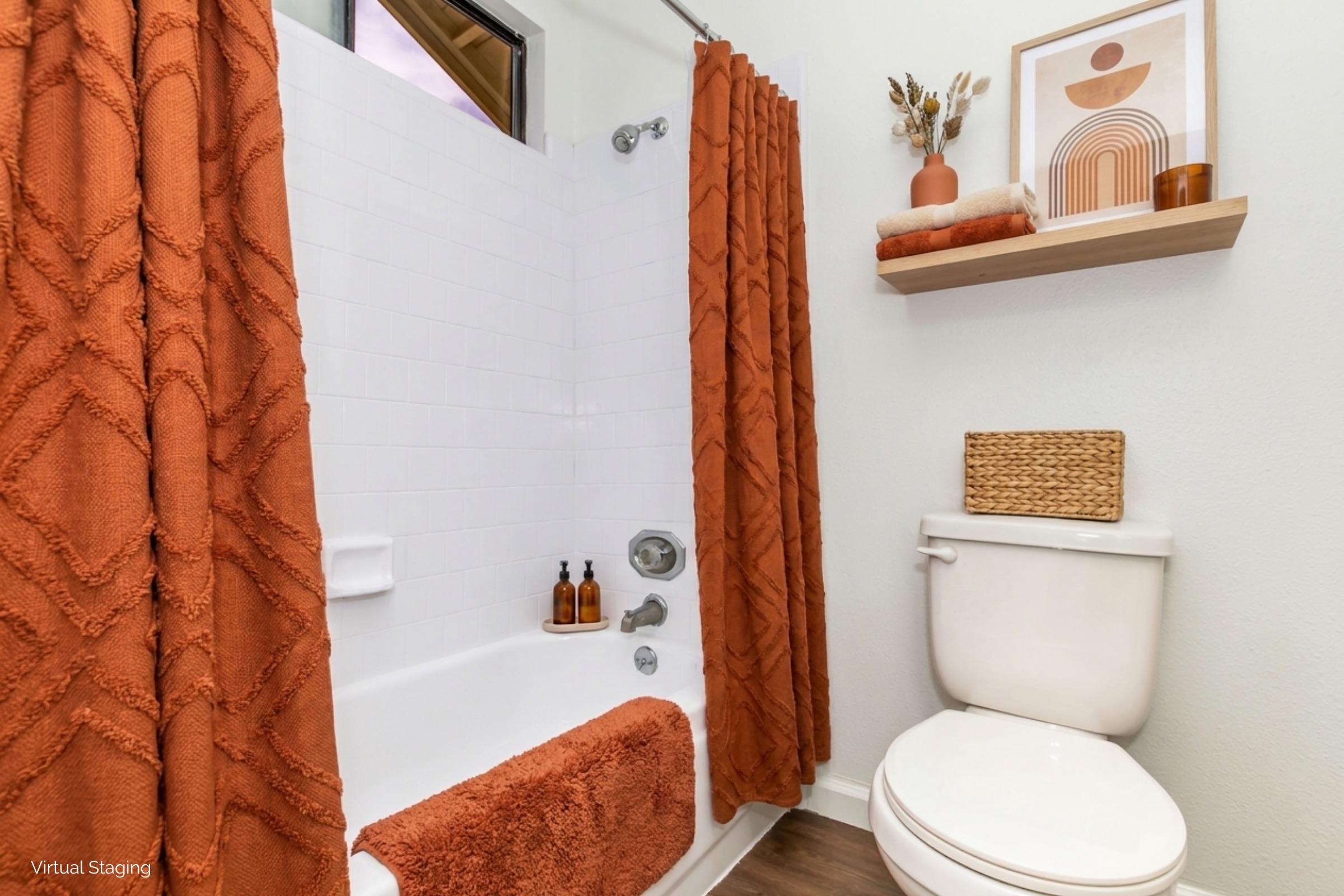 A modern bathroom featuring a white bathtub with a shower, adorned with orange textured curtains and a matching bath mat. A shelf above the toilet displays decorative items and a woven basket. The walls are painted light, and there are minimalistic decor elements for a contemporary look.