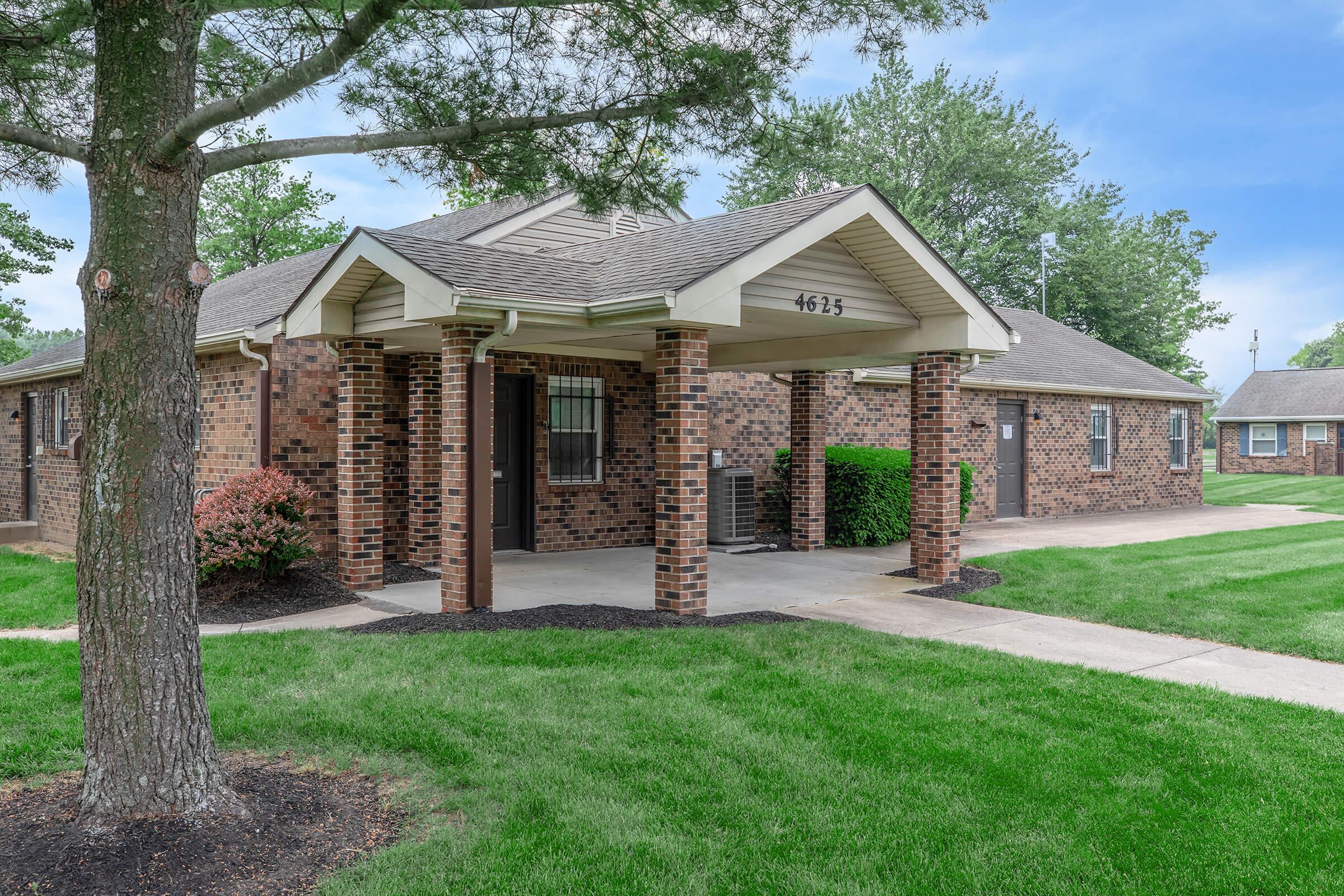 Front view of a single-story brick house with a covered porch, featuring a small landscaped yard with green grass and a few trees. The house has large windows and a wide pathway leading up to the entrance, surrounded by well-maintained shrubs and plants.
