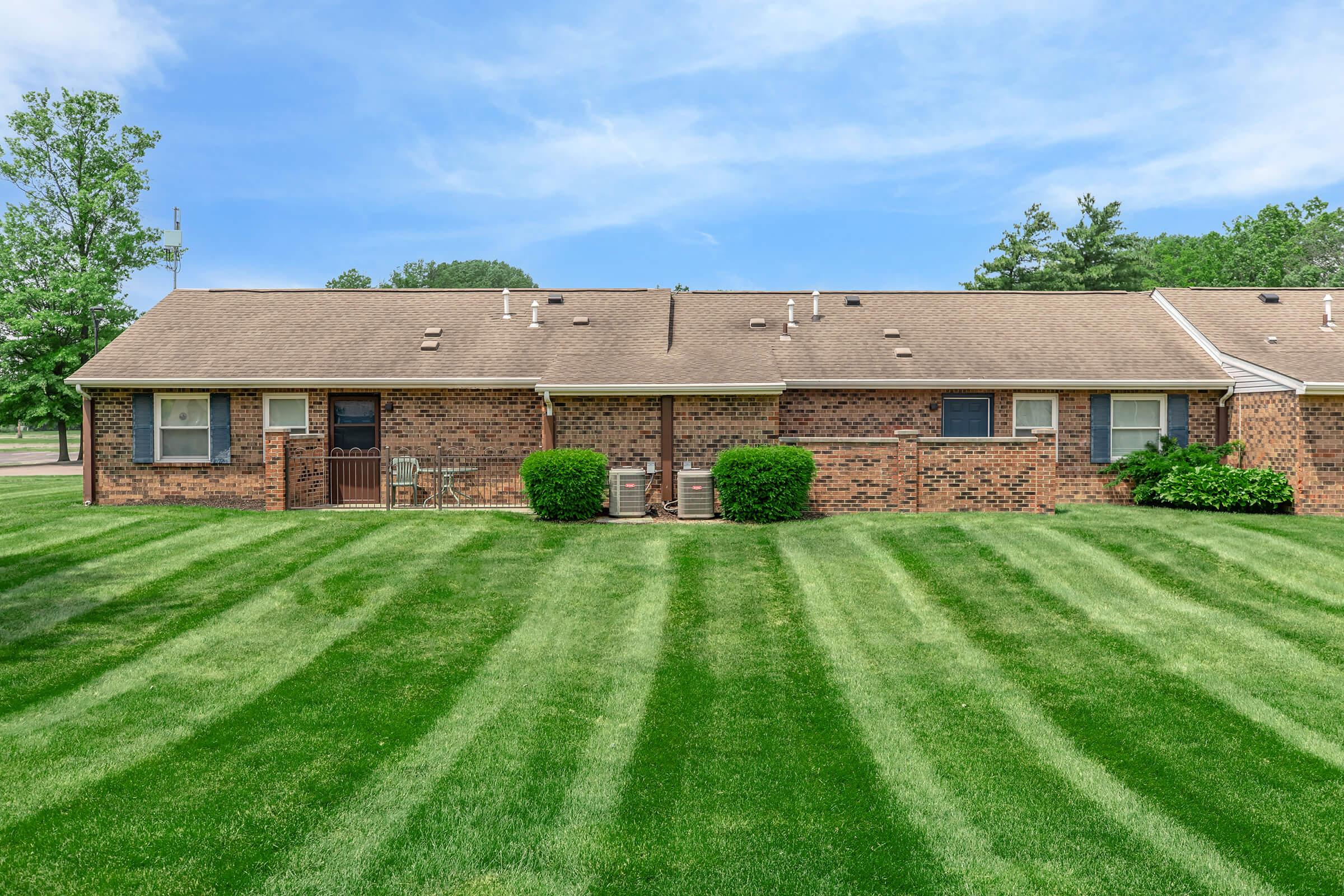 A view of a brick duplex with two units, featuring neatly trimmed green lawns with striped patterns. The sky is clear with a few clouds, and there are trees in the background. Air conditioning units are visible near the entrance of each unit, adding to the suburban atmosphere.
