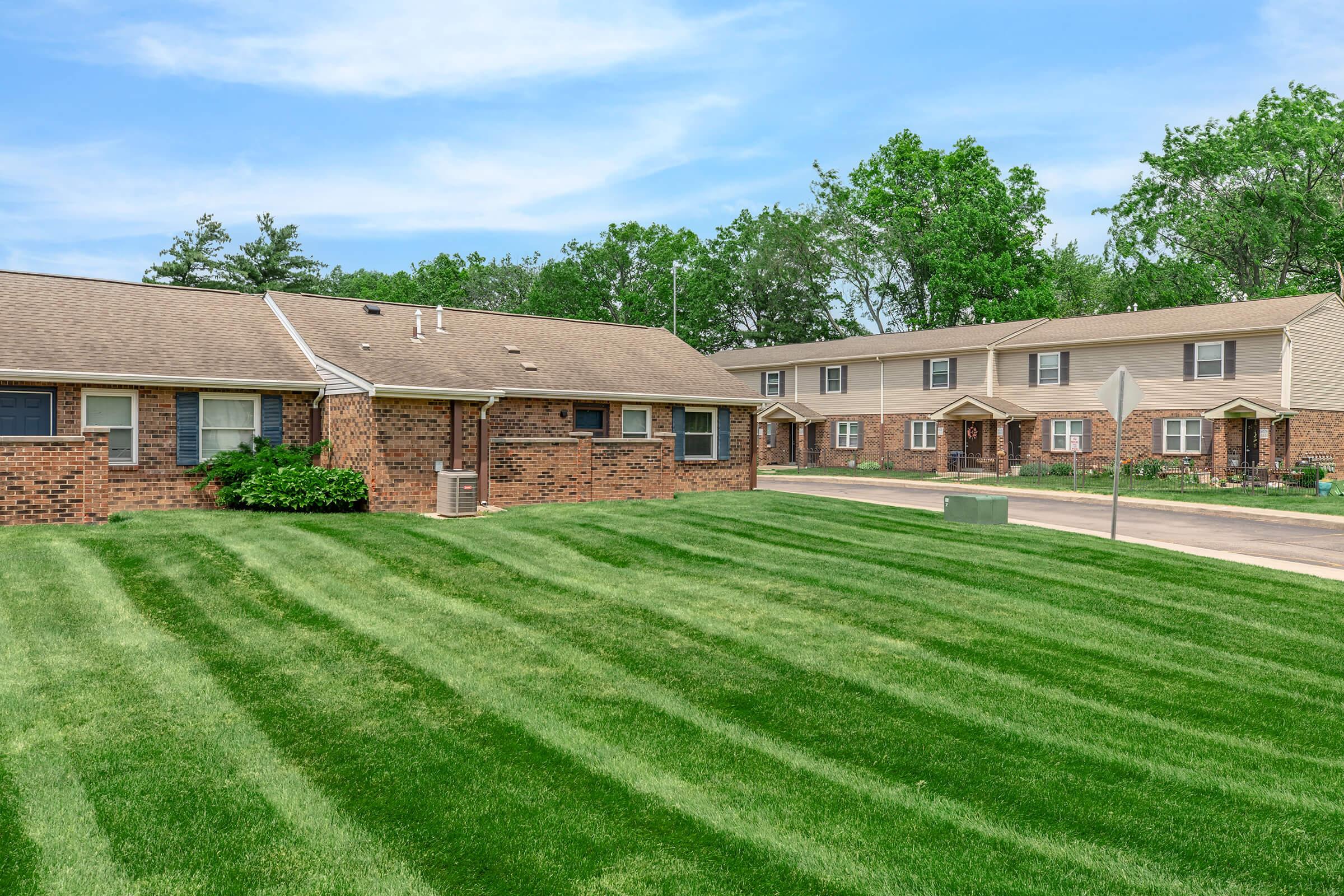 A residential area featuring brick buildings in the foreground and multi-family homes in the background. The landscape includes a well-maintained green lawn with neatly trimmed grass, trees lining the area, and a clear blue sky. The scene conveys a peaceful neighborhood atmosphere.