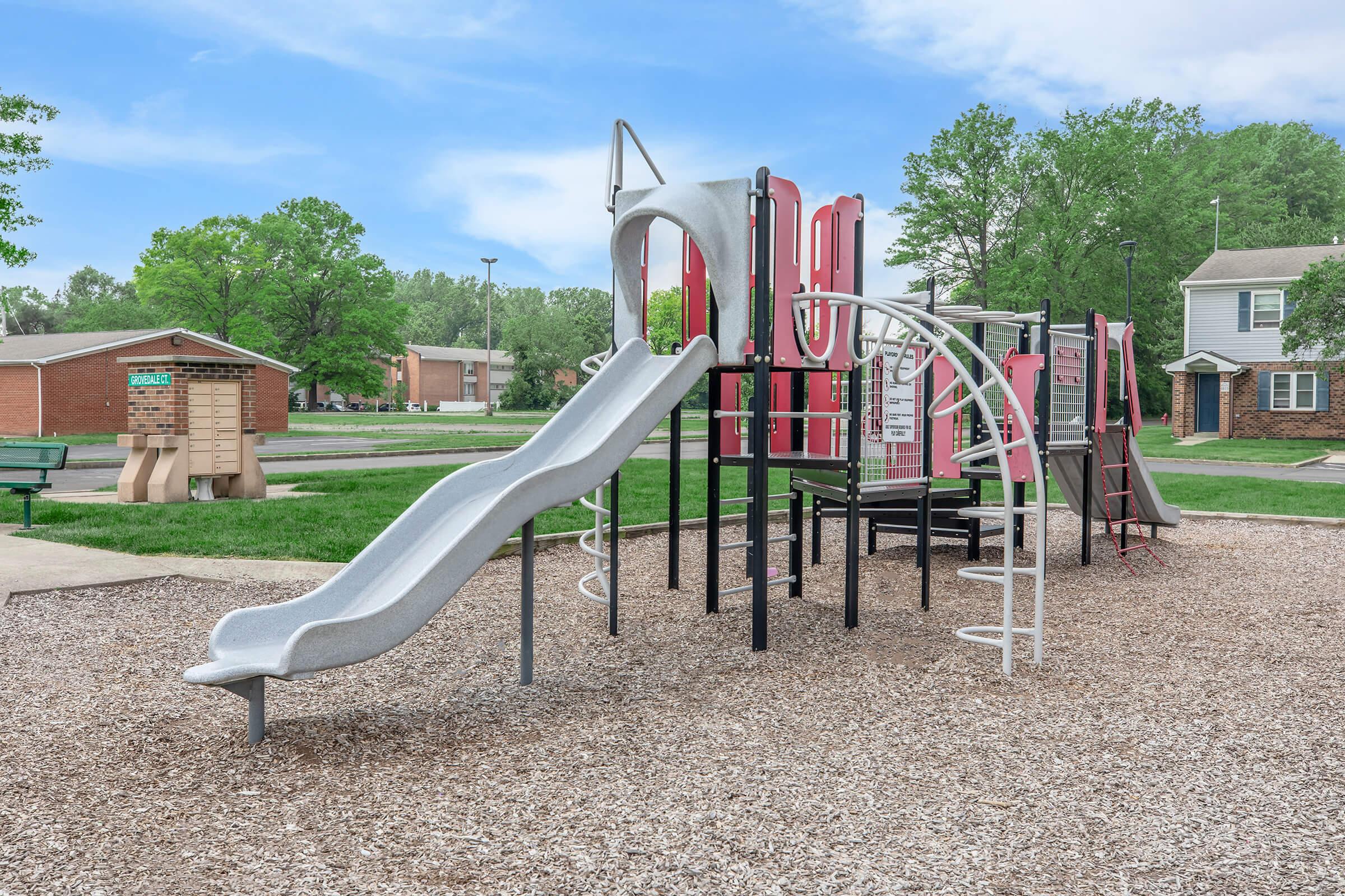 A playground featuring a gray slide, climbing apparatus, and various play structures, surrounded by soft ground covering. Green trees and a residential area are visible in the background, creating a sunny outdoor play space.