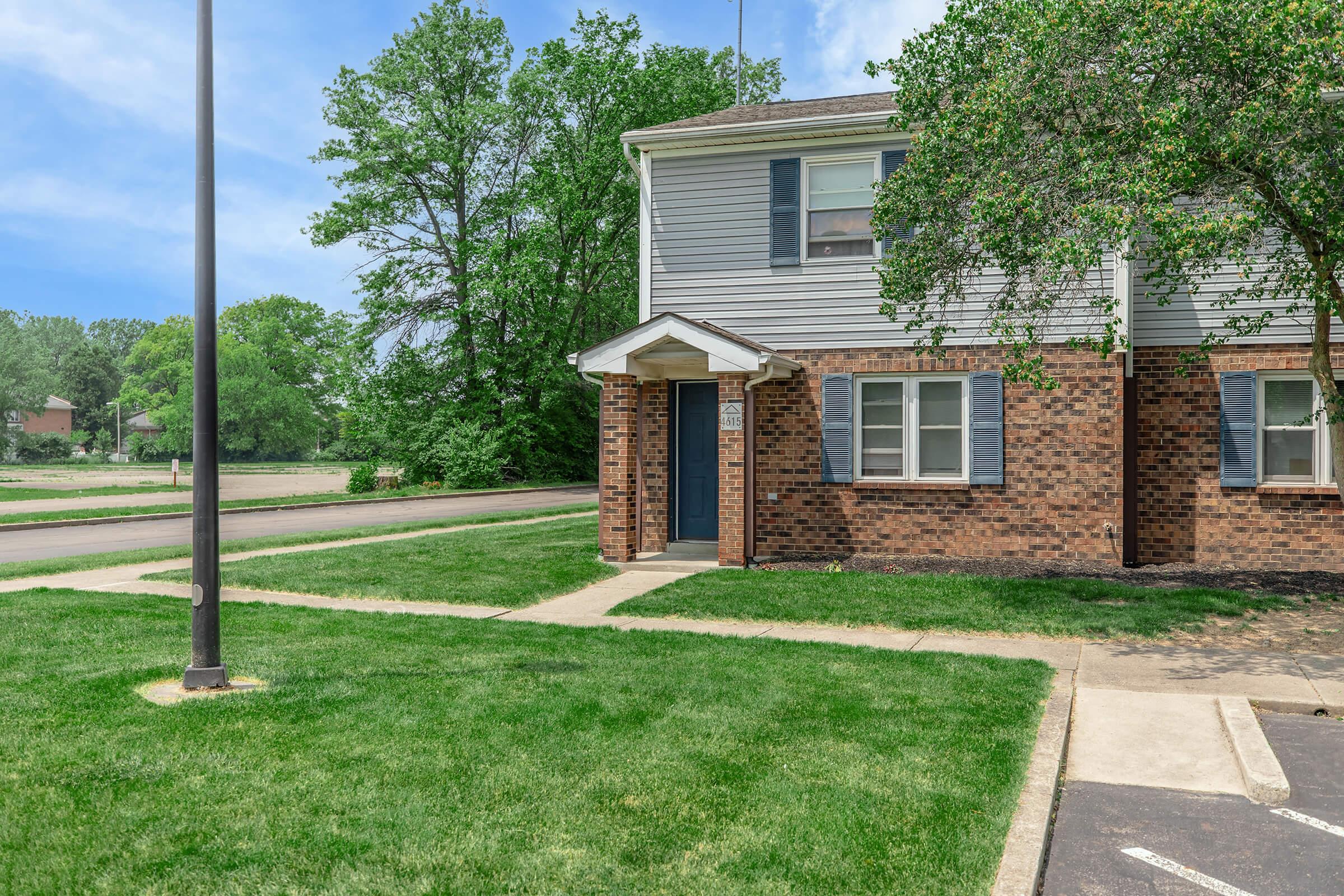 A corner of a two-story apartment building featuring a brick facade and light gray siding. The entrance has a small porch with a blue door. Surrounding the building are well-manicured lawns and trees, with a sidewalk leading to the entrance. A lamp post is visible in the foreground.
