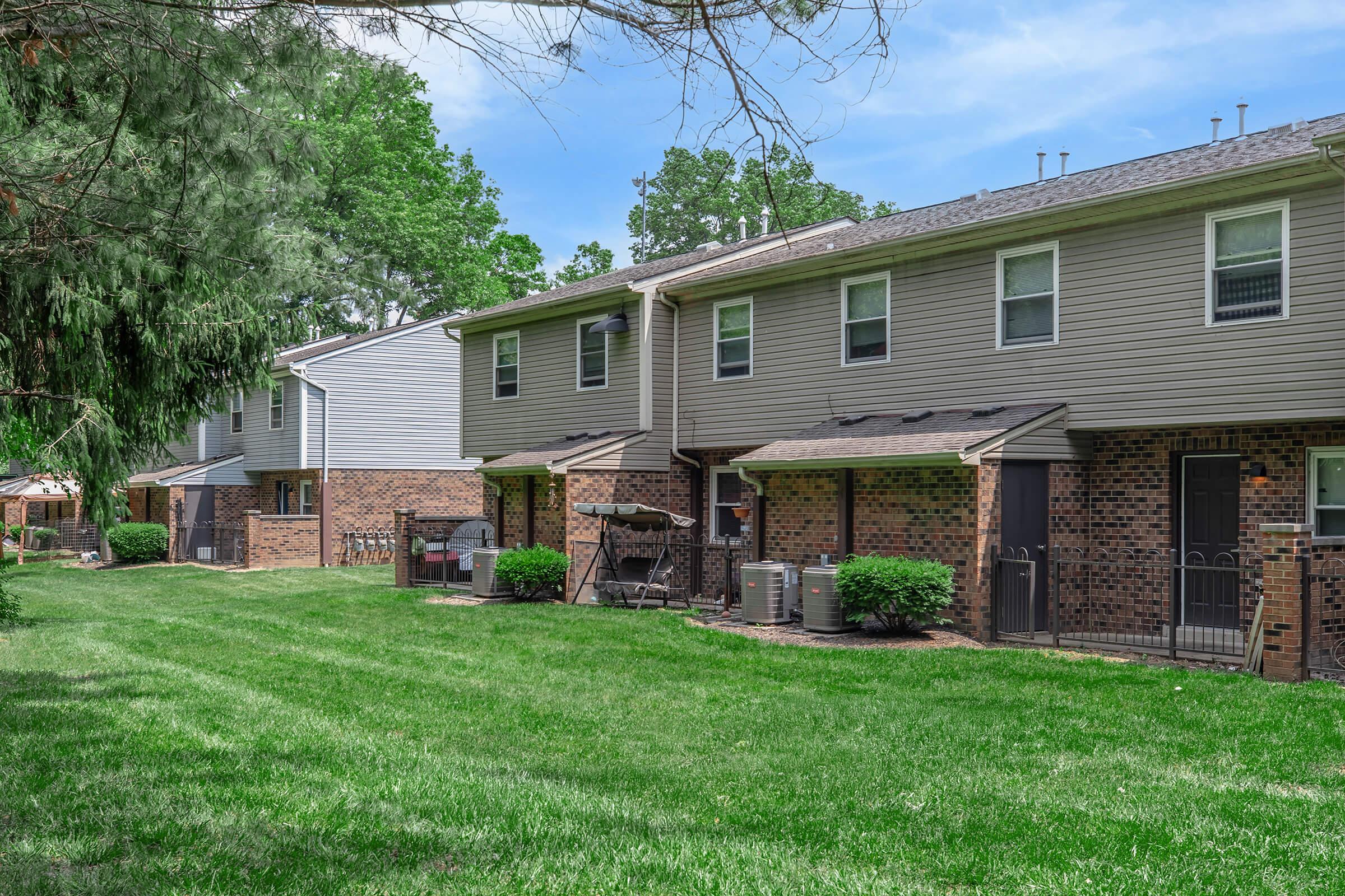 A row of townhouses with brick exteriors and grassy front lawns, set in a green landscape. Each unit features a patio area with outdoor furniture, air conditioning units, and trees providing shade. The sky is clear and sunny, enhancing the serene atmosphere of the neighborhood.