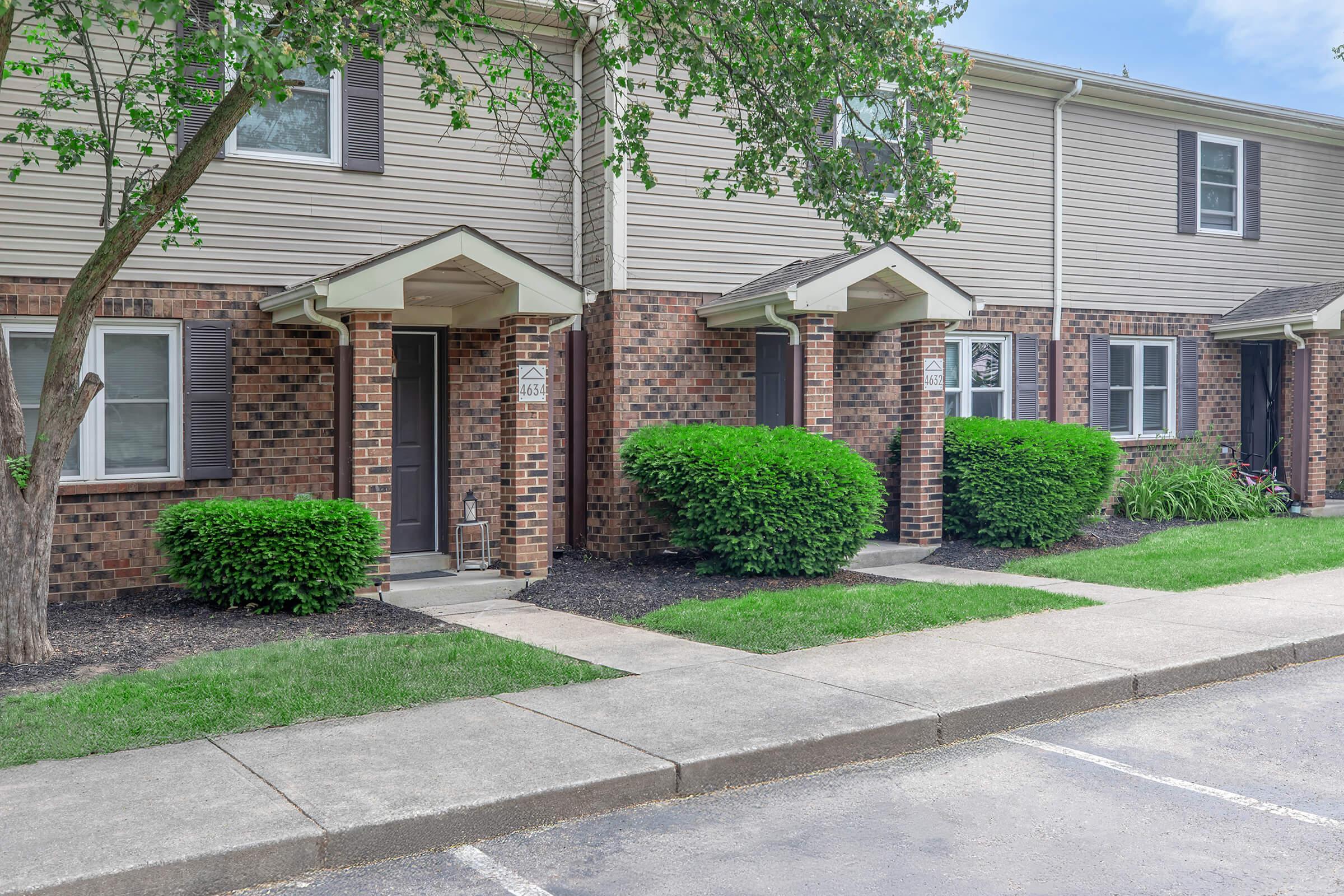 Residential building with several units, featuring brick and siding exterior. Each unit has a small covered entryway with steps leading to the door. Lush green shrubs flank the entrances, and a paved walkway runs in front of the units, alongside a parking area.