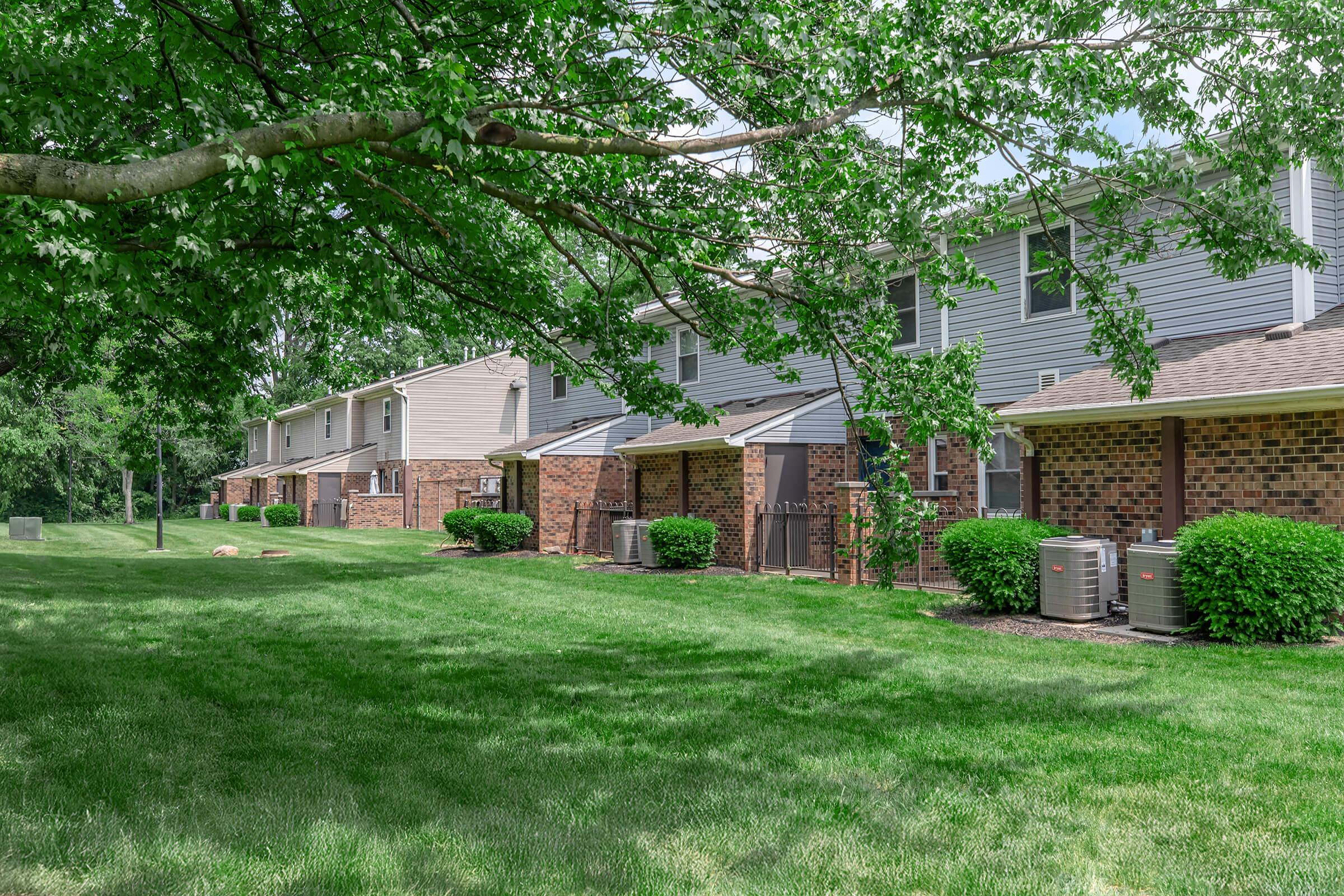 A row of two-story residential buildings with brick and siding exteriors, surrounded by lush green grass and trees. Air conditioning units are visible outside each unit, and the area appears well-maintained with landscaping features. The sky is clear, suggesting a pleasant day.
