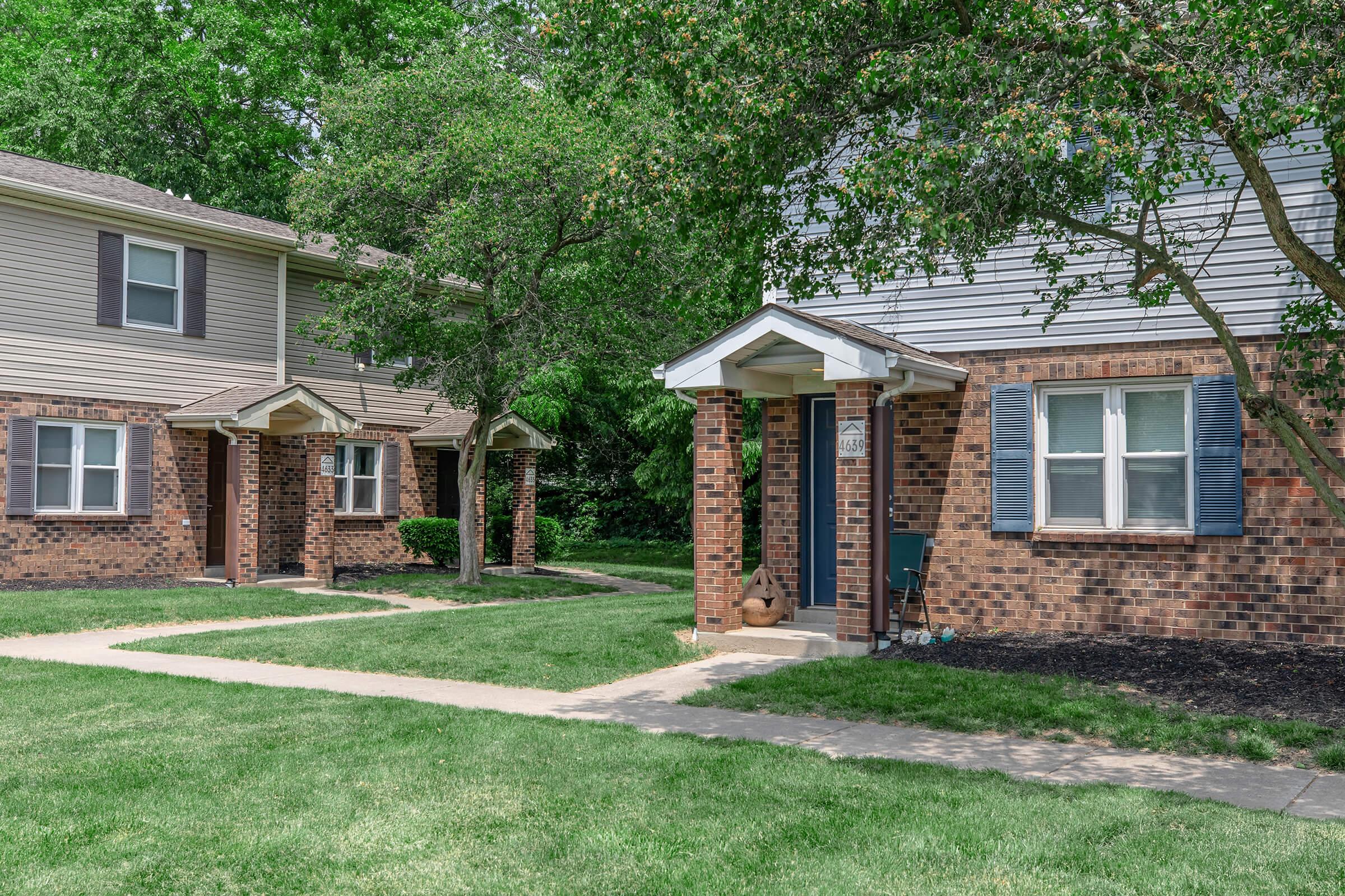 A residential area featuring two attached brick duplexes with small porches. Lush green grass surrounds the pathways leading to the front doors. Trees and shrubs add greenery to the scene, creating a peaceful suburban atmosphere.