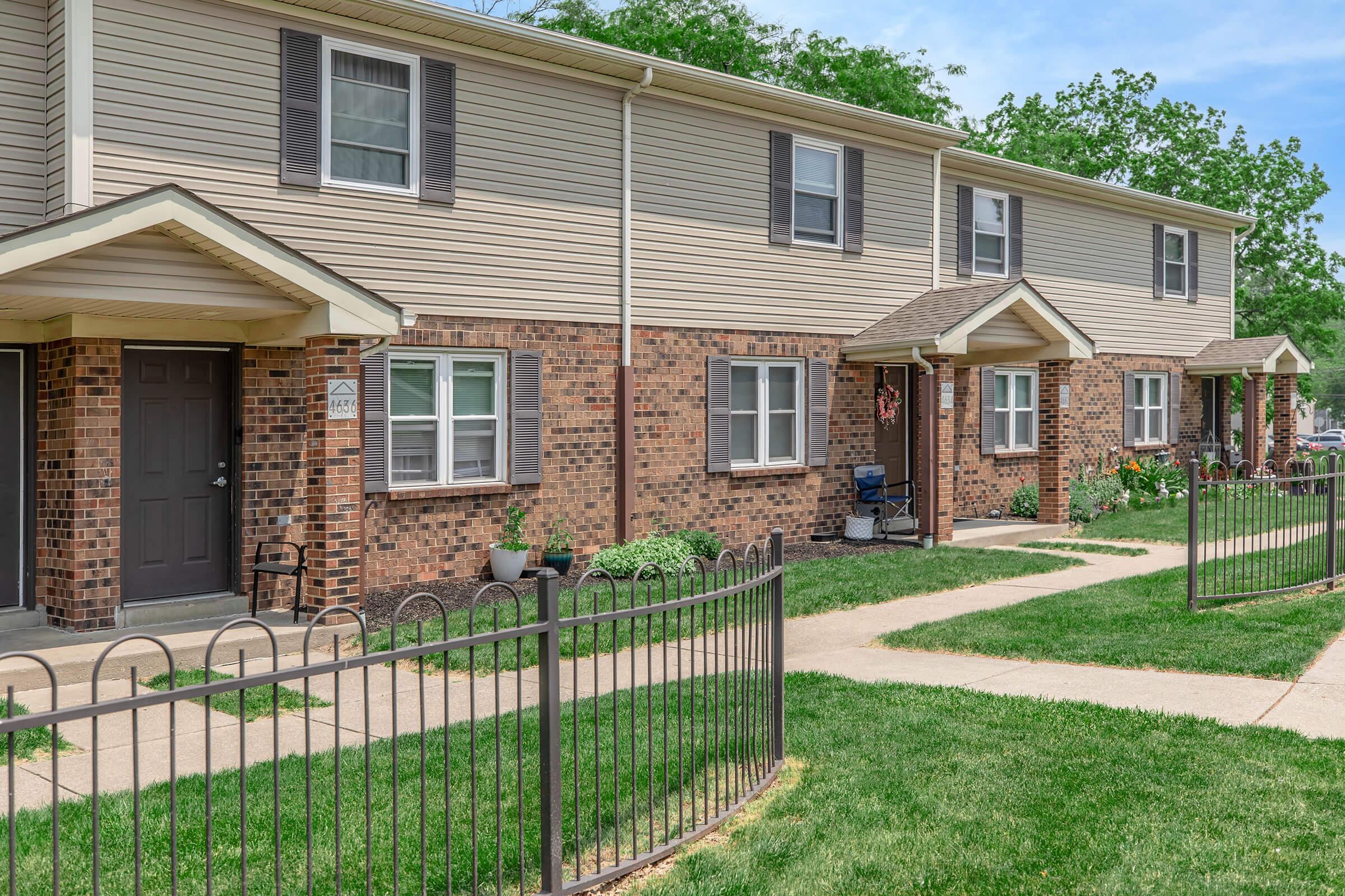 A row of townhouses featuring brick and siding exteriors. Each unit has a small porch and a well-maintained lawn with flowerbeds. Sidewalks lead to the entrances, and decorative plantings add to the curb appeal. The scene is set under a clear blue sky.