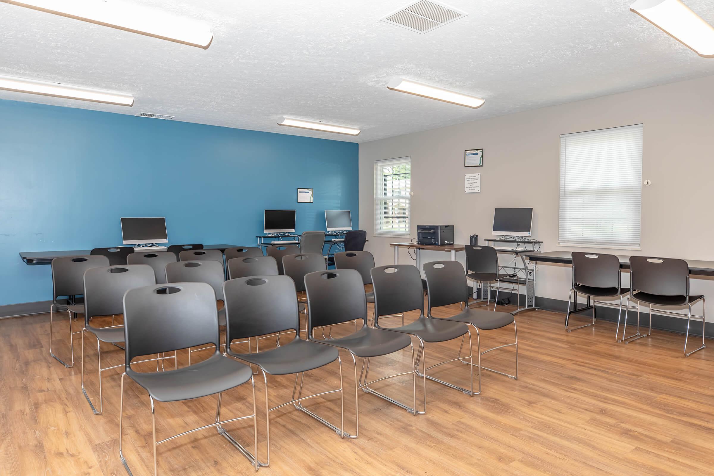 A classroom or training room featuring several black chairs arranged in rows, with a blue accent wall. There are computer desks equipped with desktop monitors, and a larger table or workstation along one wall. The room has a warm wooden floor and bright overhead lighting.