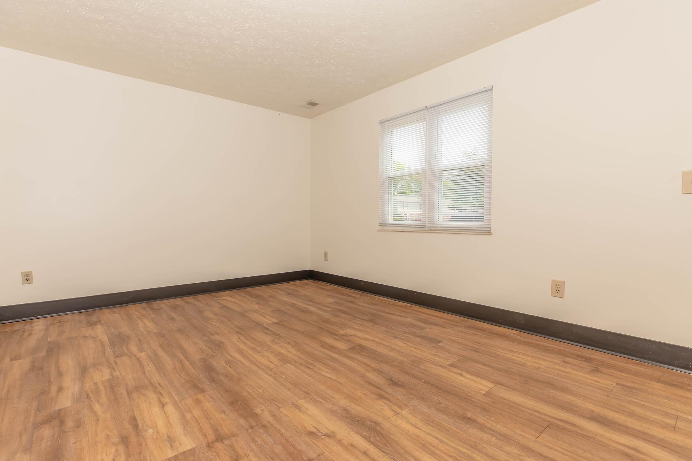 A spacious, empty room with light-colored walls and a window letting in natural light. The flooring is a warm wood finish, and the room features minimal decorative elements, creating a clean and open atmosphere.