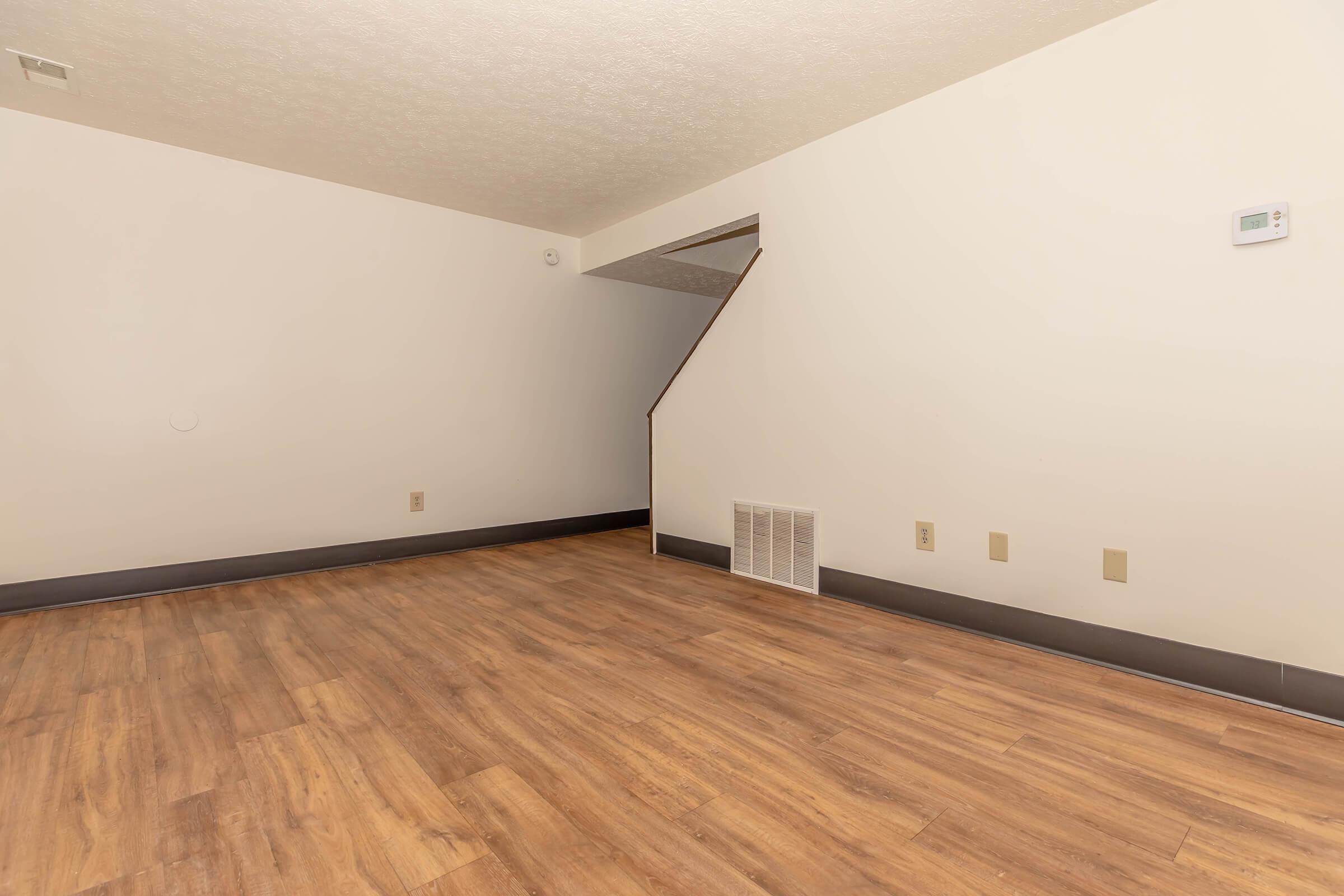 An empty room with light-colored walls and wooden flooring. There is a staircase in the background, and a heating vent on the wall. The room is well-lit and spacious, with minimal furniture or decorations.
