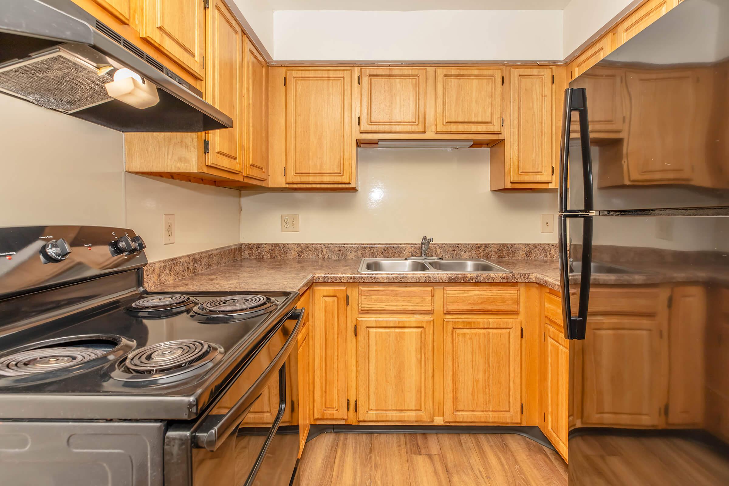A small, modern kitchen featuring wooden cabinets, a black stove with four burners, a stainless steel sink, and a black refrigerator. The countertops are a dark stone pattern, and the flooring has a wood-like appearance. The space is well-lit with natural light.