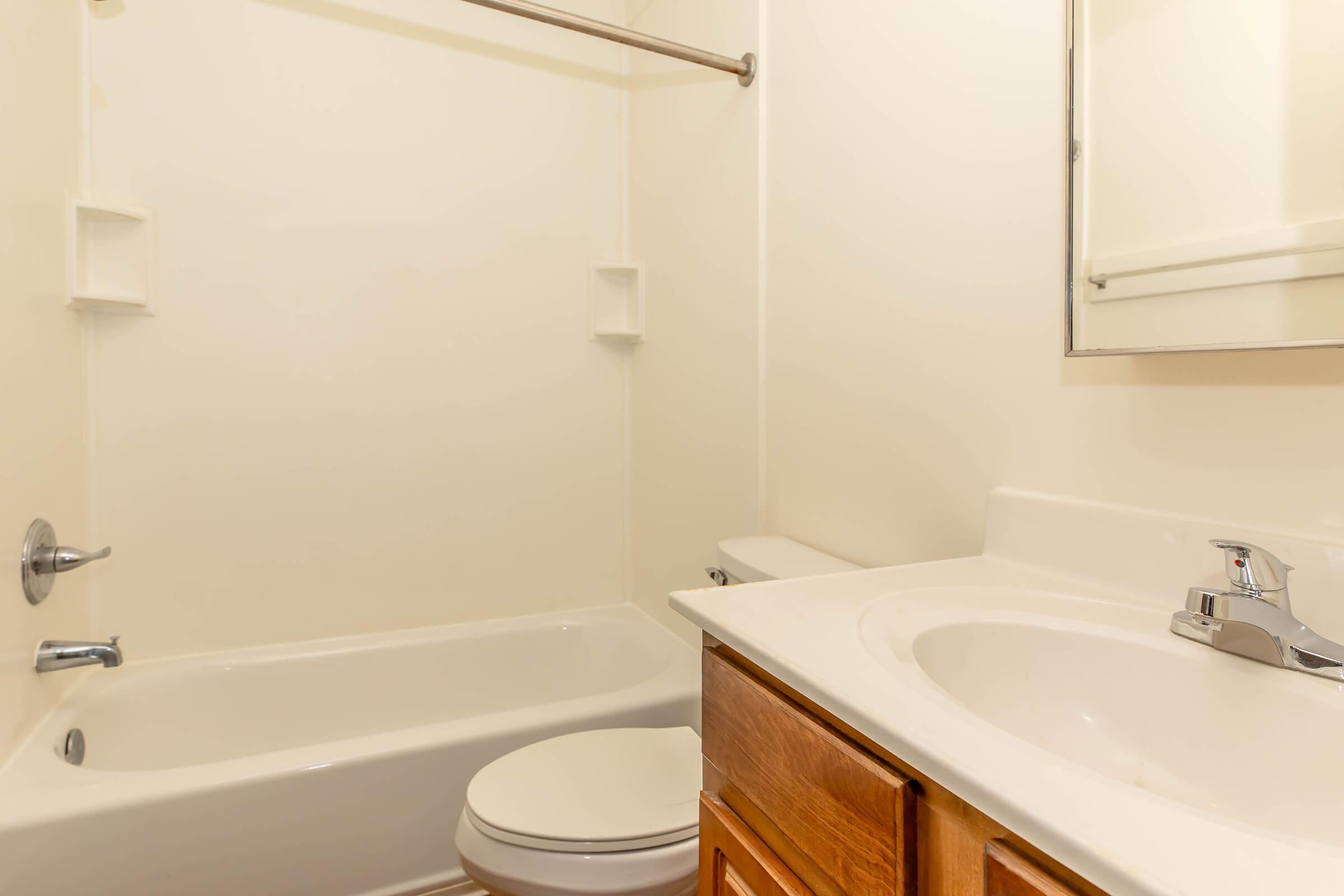 A clean, simple bathroom featuring a bathtub with a shower rod, a toilet, and a sink with a mirror above it. The walls and tiles are painted in a light color, creating a bright and spacious atmosphere. Wooden cabinetry adds a touch of warmth to the space.