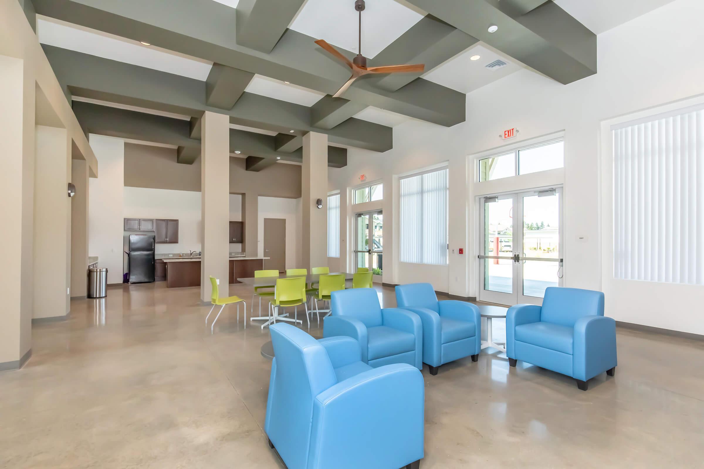 A spacious, modern indoor lounge area featuring blue armchairs, round dining table with green chairs, and a kitchen area in the background. Large windows provide natural light, and a ceiling fan hangs above. The decor emphasizes a sleek and contemporary design.