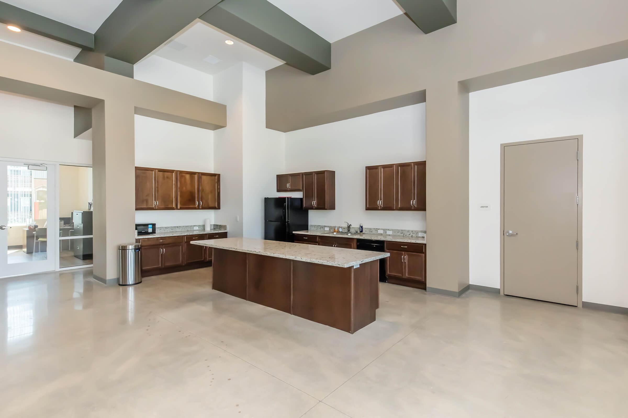Spacious modern kitchen with high ceilings, featuring dark wood cabinets, a large central island with a granite countertop, and stainless steel appliances. The floor is polished and there is a doorway leading to another room. Natural light enters through large windows.