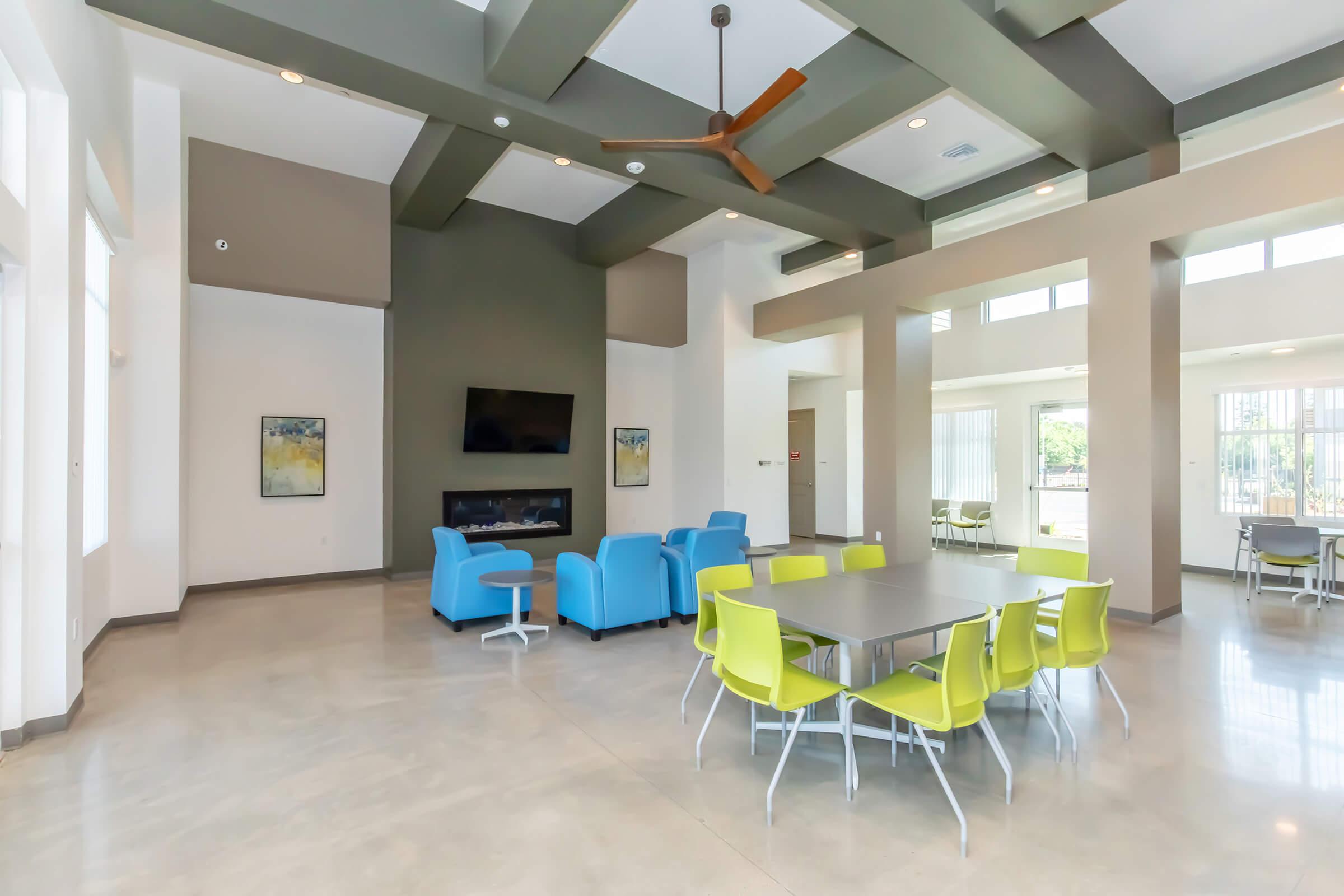 Modern communal space featuring a seating area with blue chairs and a table surrounded by bright yellow chairs. Thereโs a flat-screen TV mounted on a dark wall and a fireplace, with large windows allowing natural light to fill the room. The floor is polished concrete, enhancing the contemporary design.