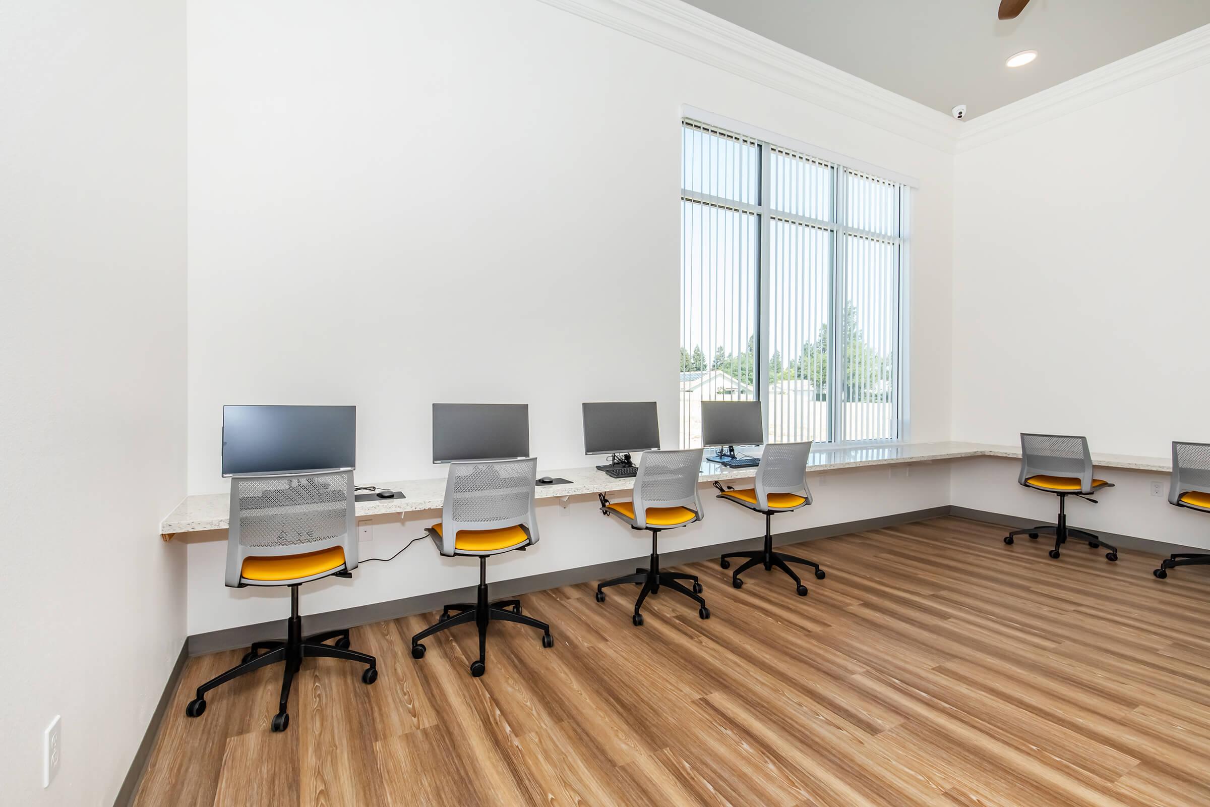 A spacious, well-lit room featuring five computer desks with modern, minimalist design. Each desk has a computer monitor and is accompanied by a yellow and gray ergonomic chair. Large windows let in natural light, illuminating the light-colored walls and wooden flooring.