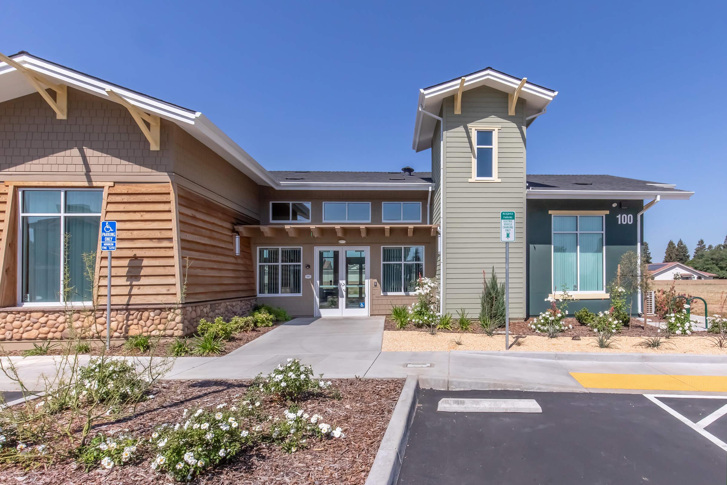 Modern building exterior featuring a combination of wooden and horizontal siding. The structure has large windows, a prominent entrance, and landscaping with plants and flowers. A parking lot is visible in front, with designated accessible parking spaces. Clear blue sky in the background.