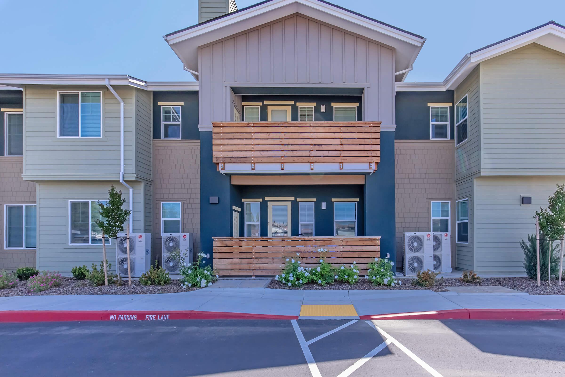 Exterior view of a modern apartment building with two balconies, surrounded by landscaping. The building features a mix of siding materials in neutral tones and has several air conditioning units on the ground floor. A red no-parking zone is marked in front of the building.