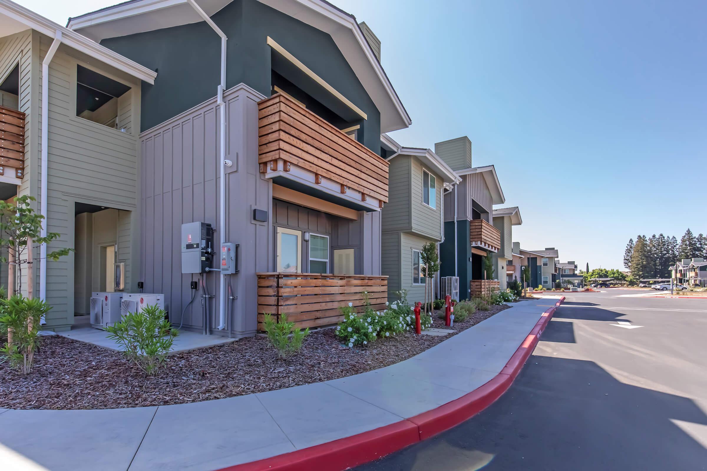 Modern multi-unit residential buildings featuring balconies, landscaped areas, and paved walkways. The architecture includes a mix of colors and materials, with a clear blue sky overhead. Paths are designed for accessibility, leading to individual units in a landscaped environment.