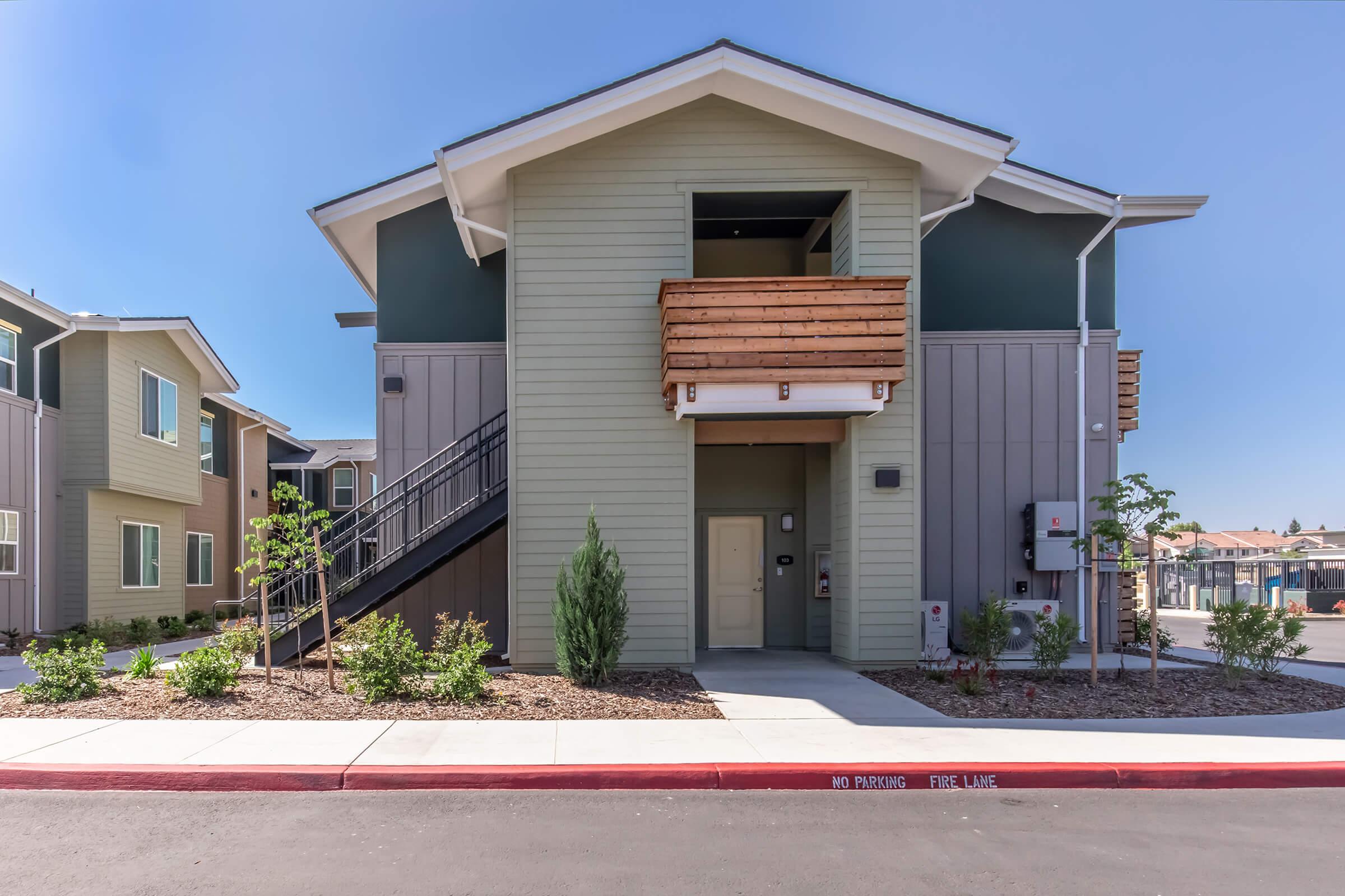 Urban residential building with a modern design featuring a two-story structure. It has a staircase on the left leading to the upper level, a covered balcony, and a front door. Landscaping includes small shrubs and trees, with a no-parking sign on the red curb near the entrance. Clear blue sky above.