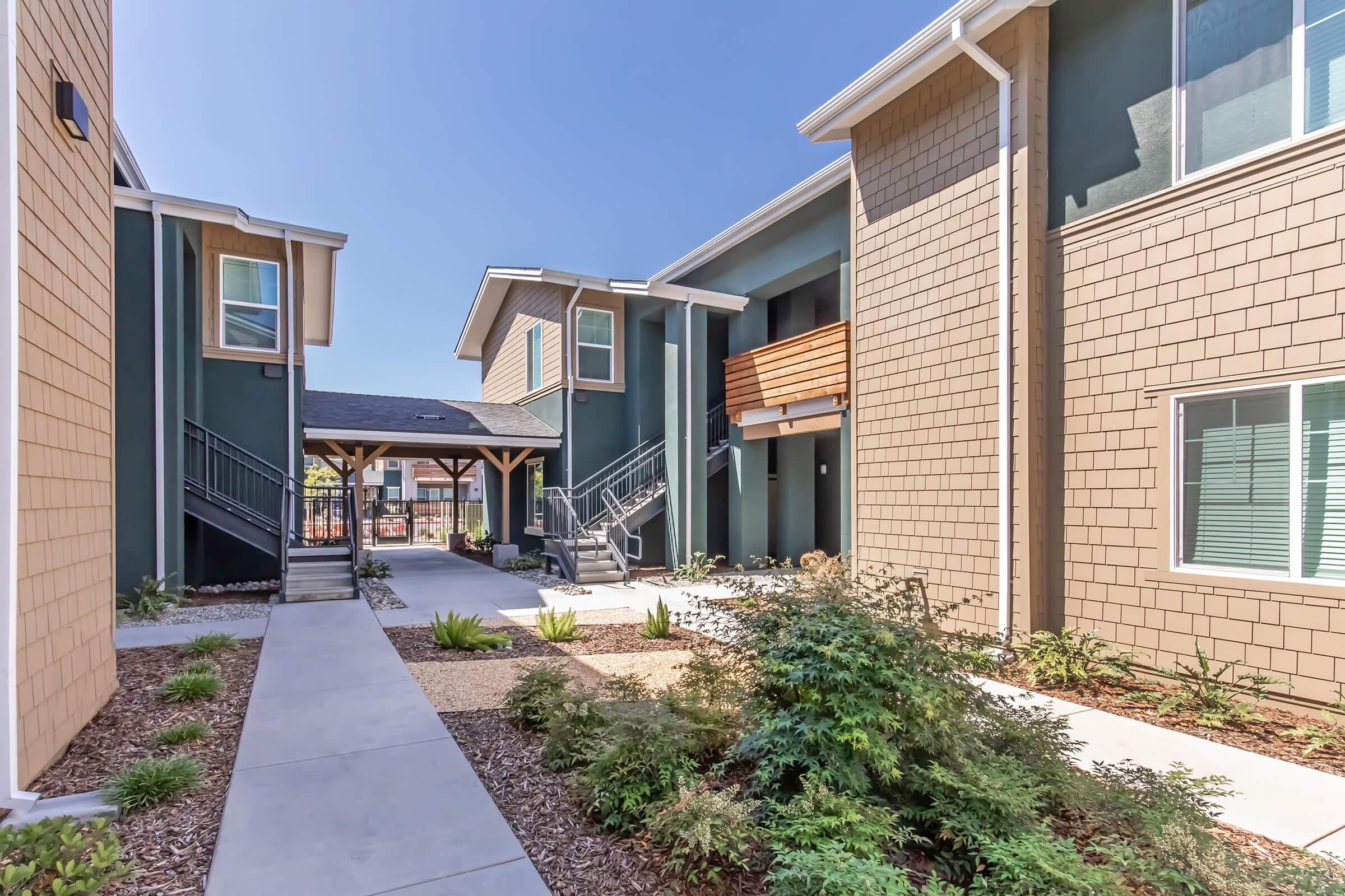 A landscaped courtyard with a pathway surrounded by two modern multi-story buildings. The buildings feature a mix of green and tan siding, with some balconies visible. Lush greenery and small plants line the walkway, and a clear blue sky is seen above.