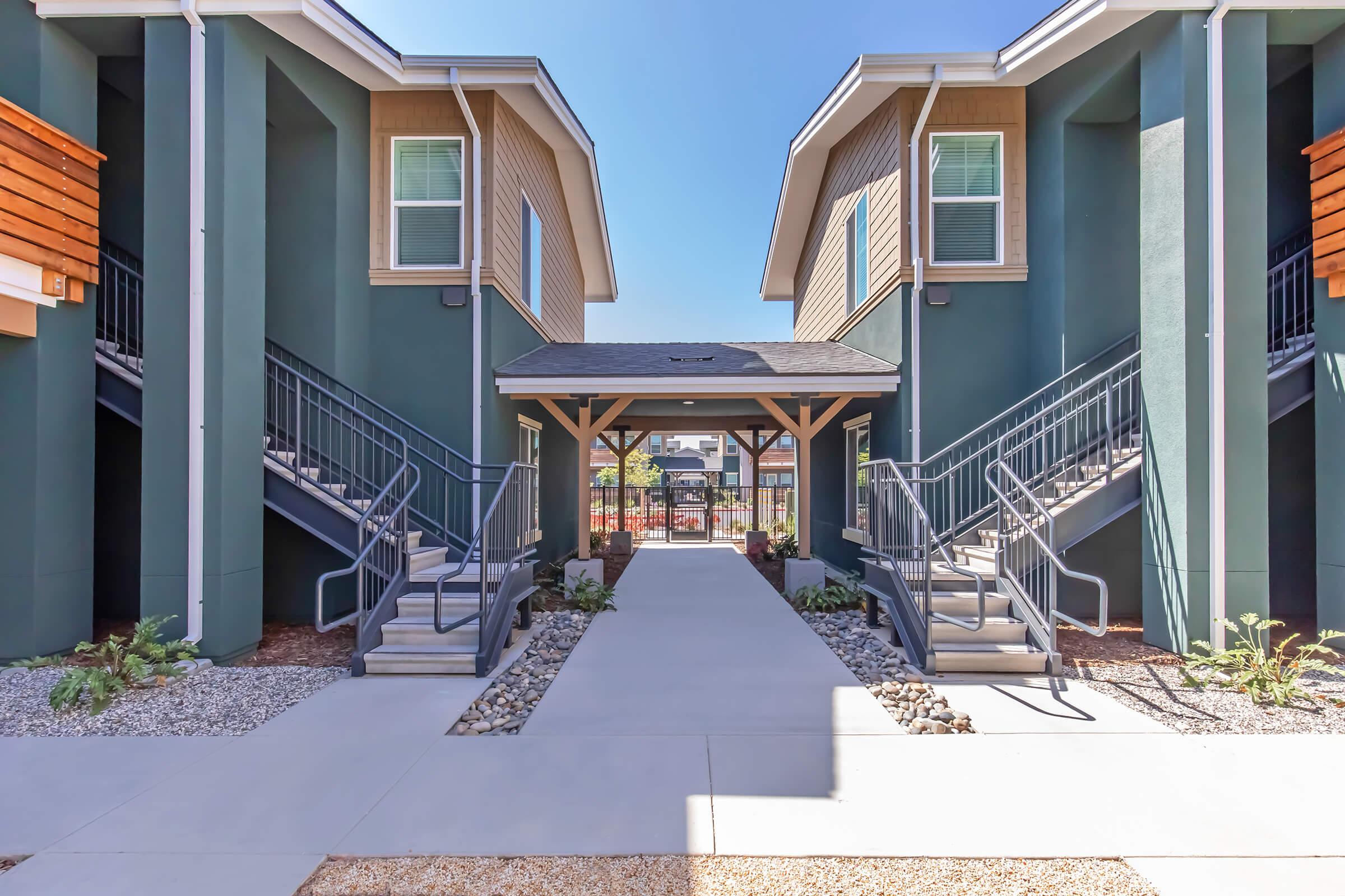 View of a landscaped pathway between two modern apartment buildings. The path features concrete walkways, decorative rocks, and greenery, with staircases leading to the upper levels of the buildings. The scene is bright and inviting, set against a clear blue sky.