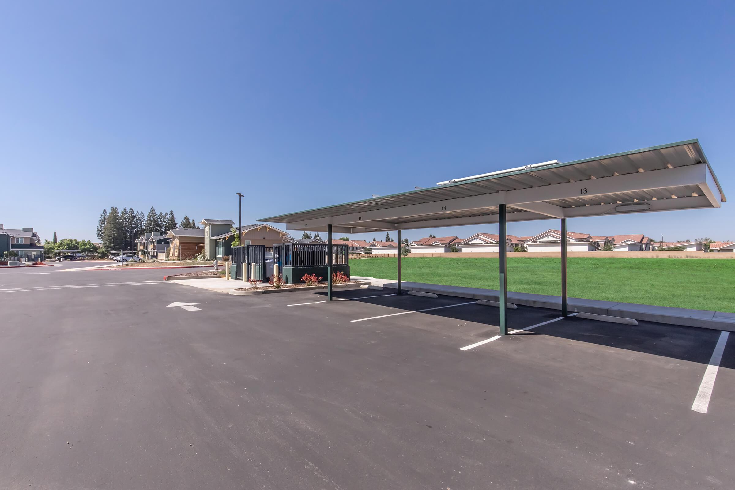 A wide view of an empty parking lot with a shaded carport structure. In the background, there are residential buildings and a green lawn. The sky is clear with no clouds, indicating a sunny day. The parking spaces are clearly marked, and there is minimal traffic or activity in the area.