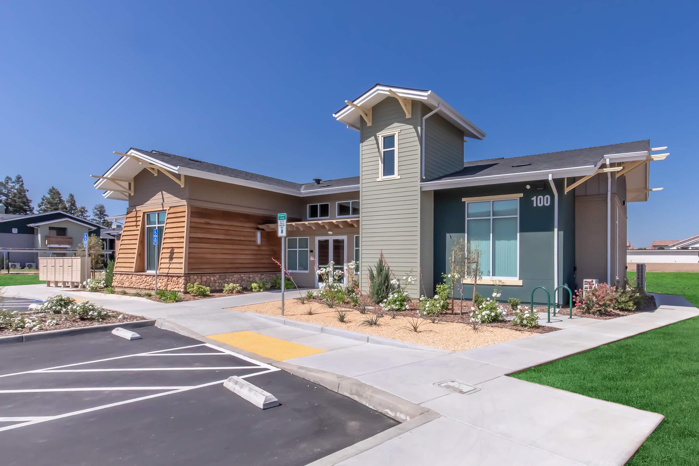Modern two-story building with a combination of wood and green exterior. Features large windows, a landscaped front yard with shrubs, and a parking lot with marked spaces. The sky is clear and blue, indicating a sunny day.