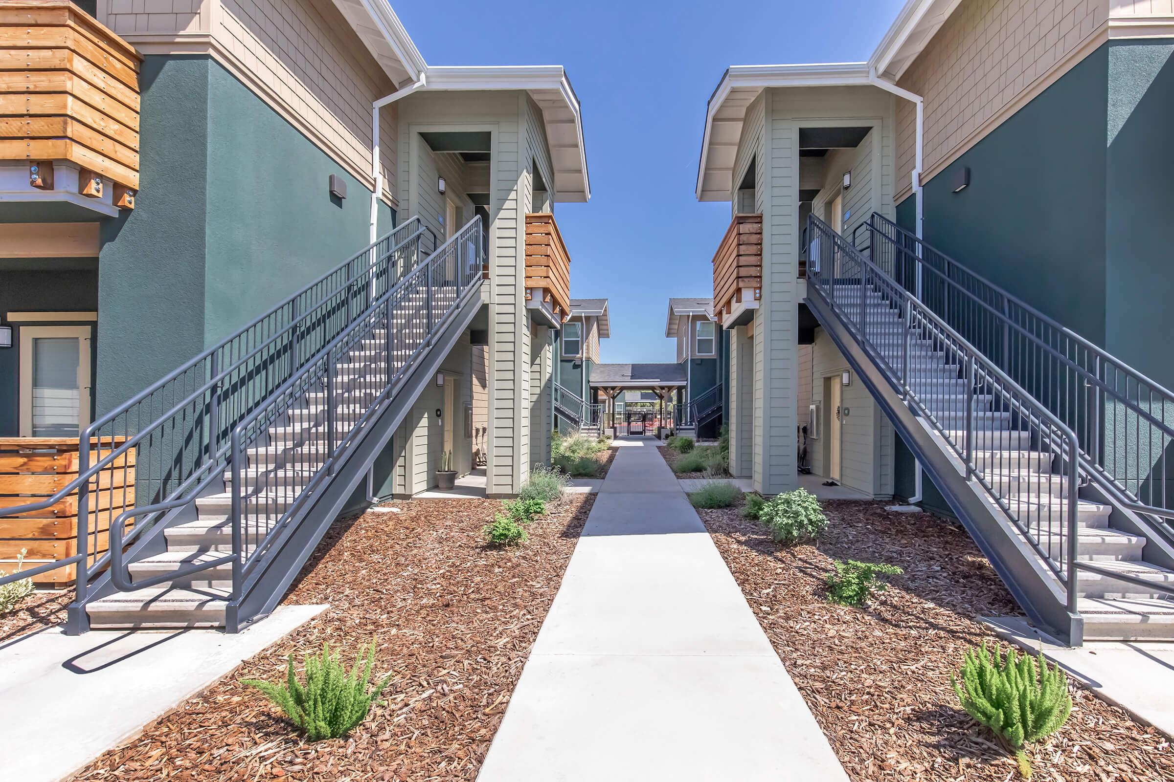 A view of a modern apartment complex featuring two sets of staircases leading to upper units. A wide concrete pathway runs between the buildings, lined with greenery and mulch, under a clear blue sky. The architecture combines various materials and colors, creating a contemporary aesthetic.