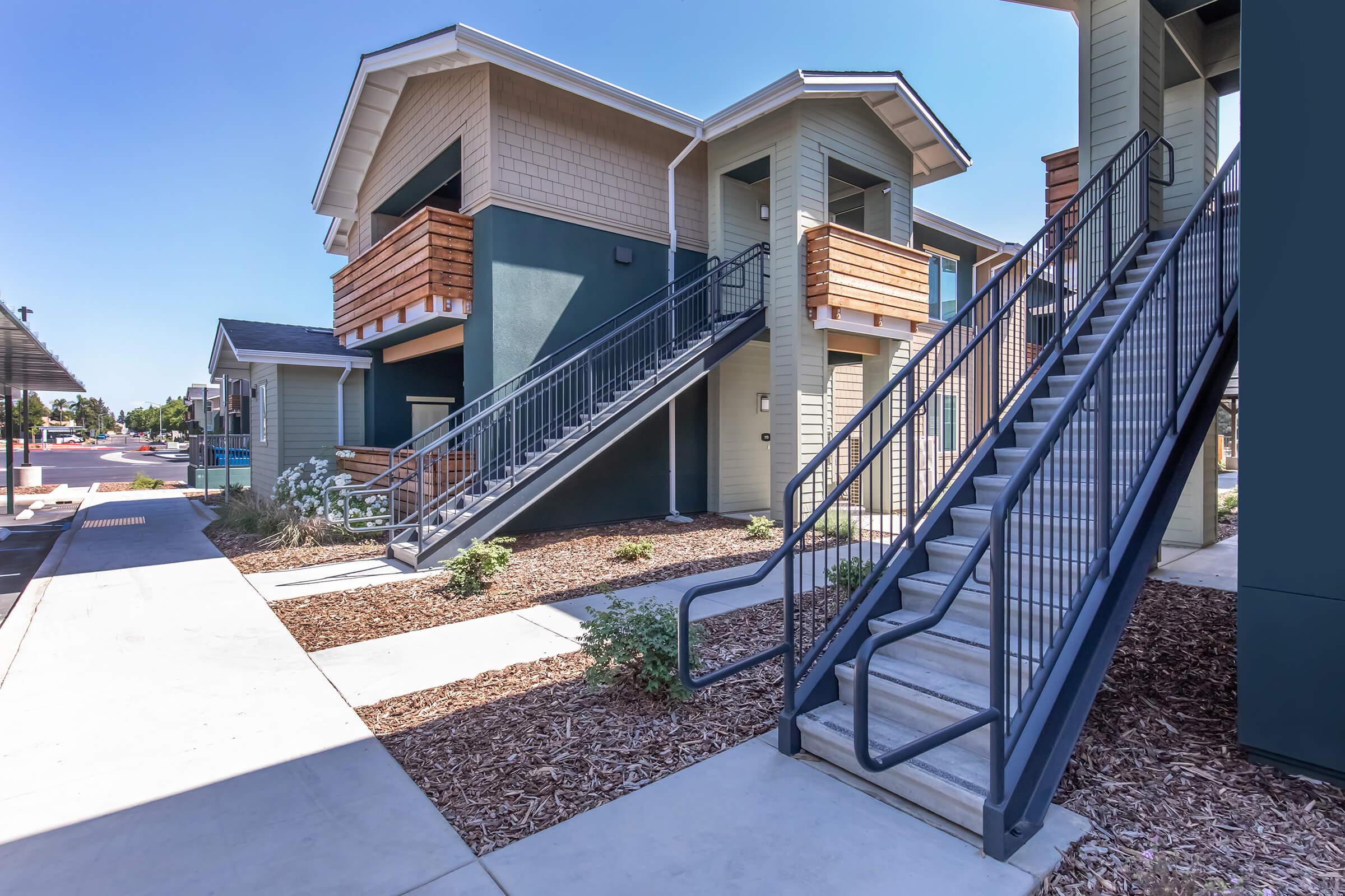 Two-story residential building with a modern design, featuring a mix of wood and green siding. Sturdy metal staircases lead to second-floor entrances, flanked by landscaped areas with mulch and small plants. A clear blue sky is visible above, enhancing the inviting atmosphere of the complex.