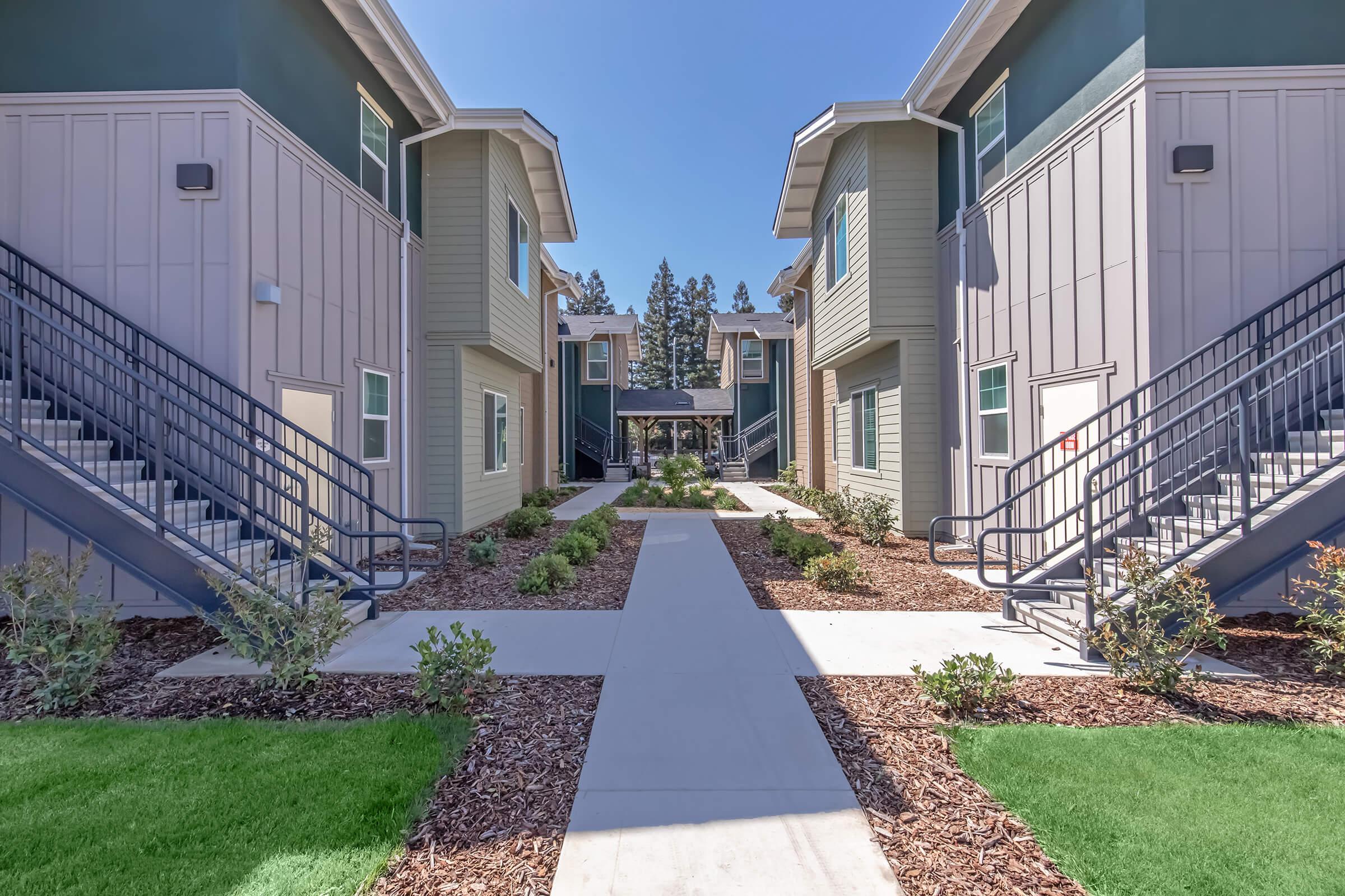 A view of a modern apartment complex with two-story buildings on either side of a central walkway. Lush green landscaping and shrubs line the pathway, leading to a courtyard area. Sturdy stairs provide access to the upper levels of the buildings, under a clear blue sky.