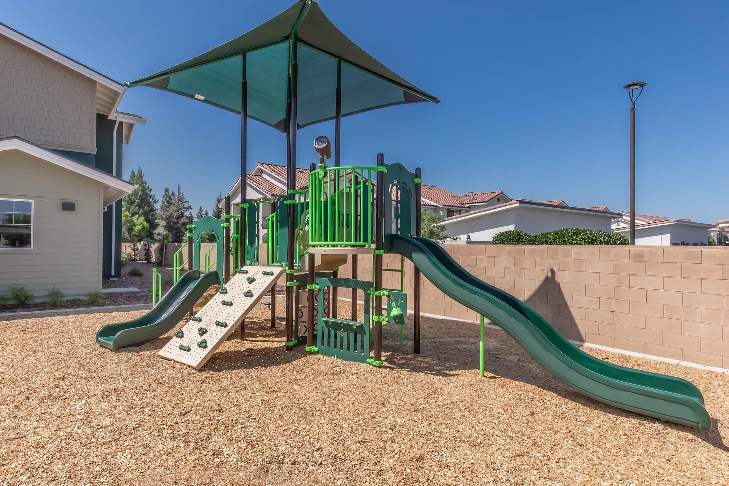 A green playground structure with slides, climbing features, and shaded areas, situated in a yard with wood chips. Residential houses can be seen in the background under a clear blue sky.