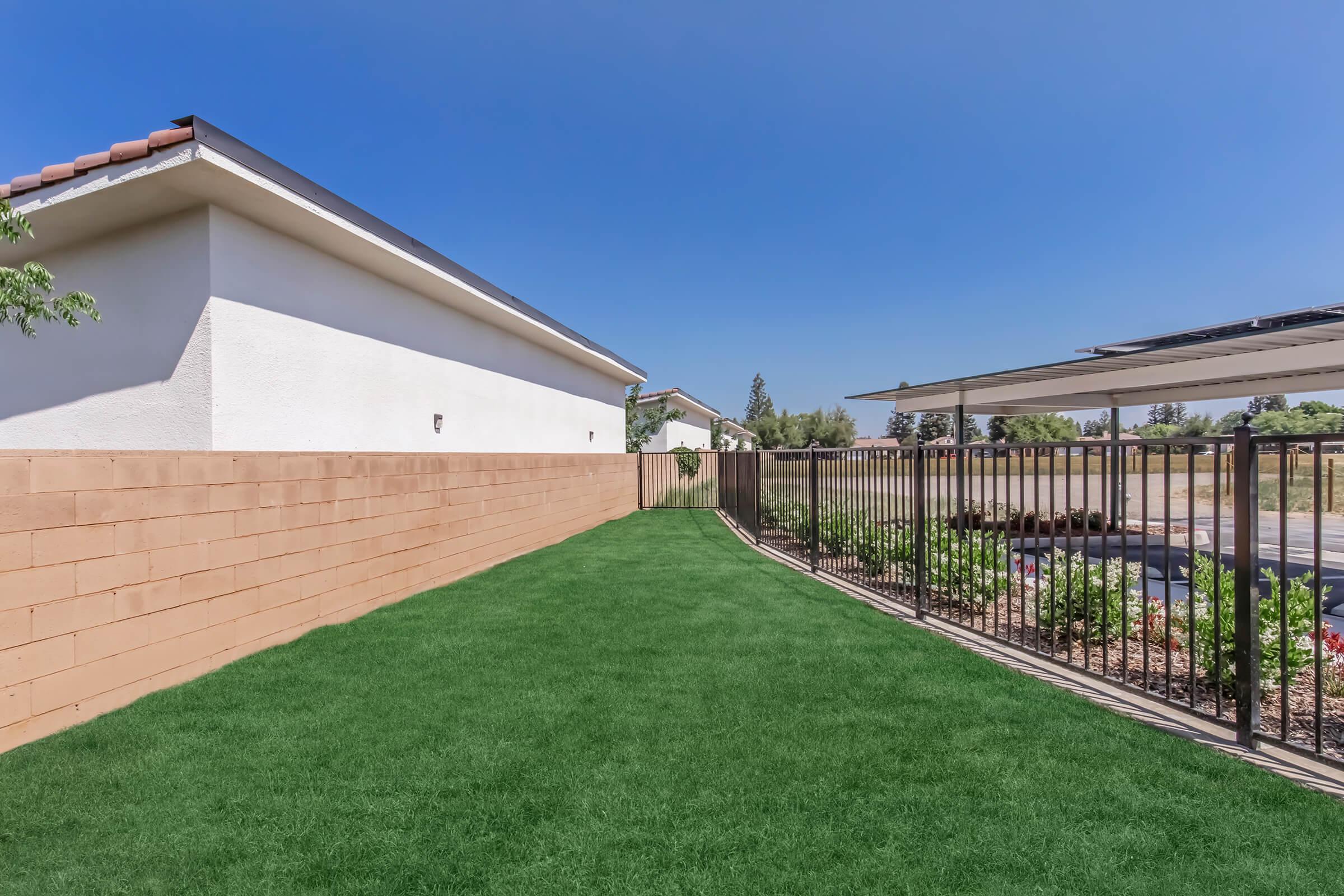 A well-maintained backyard featuring an expanse of green artificial grass, bordered by a beige wall on one side and a black metal fence on the other. A patio area with solar panels is visible in the background, along with landscaped plants and bushes. The sky is clear and blue.