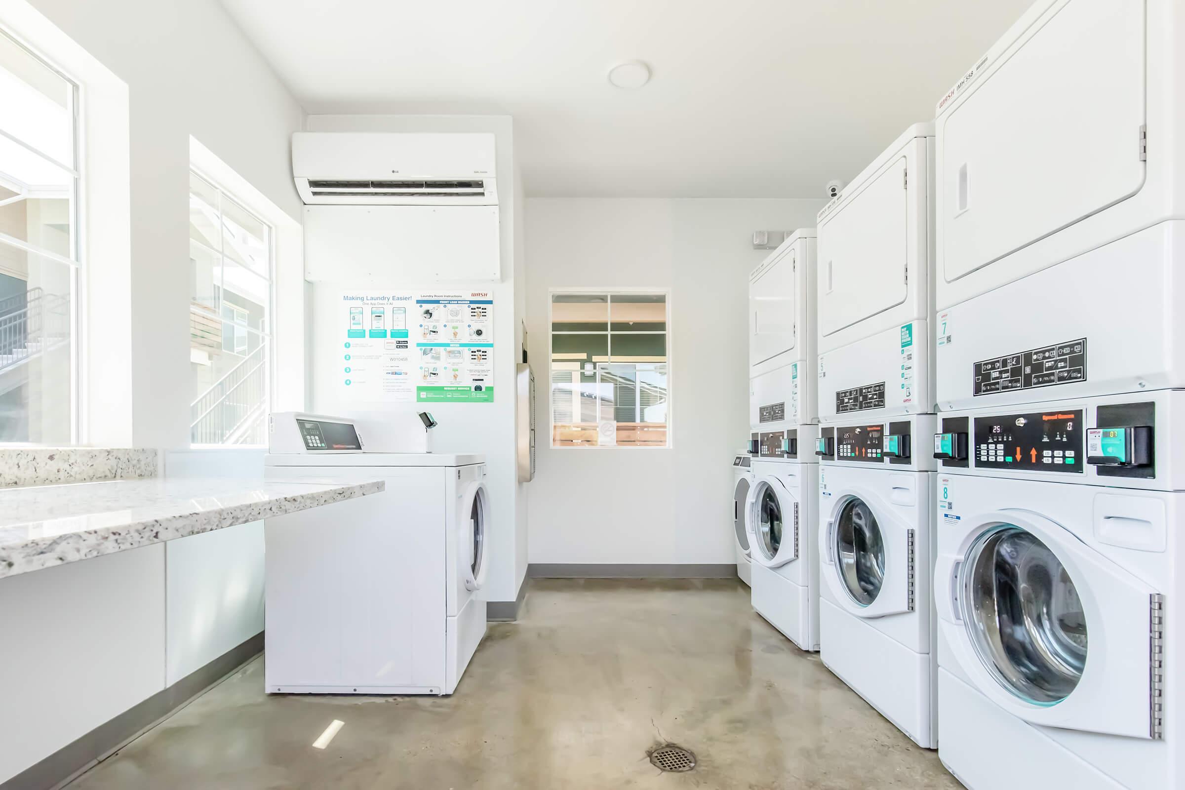A bright and modern laundry room featuring several stacked washing machines and dryers against a clean, light wall. There's a counter on the left for folding clothes, and large windows provide natural light. A bulletin board with laundry instructions is visible on the wall.