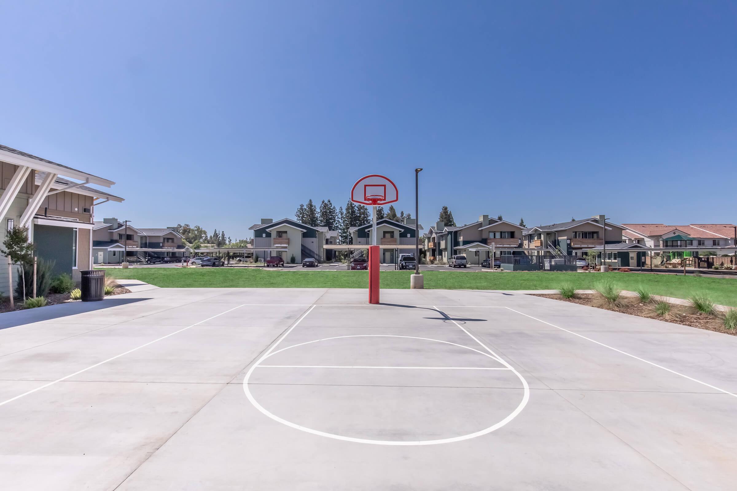 A bright outdoor basketball court featuring a red hoop and clear blue skies. The court is surrounded by well-maintained grass and residential buildings in the background, showcasing a sunny day with no people present.